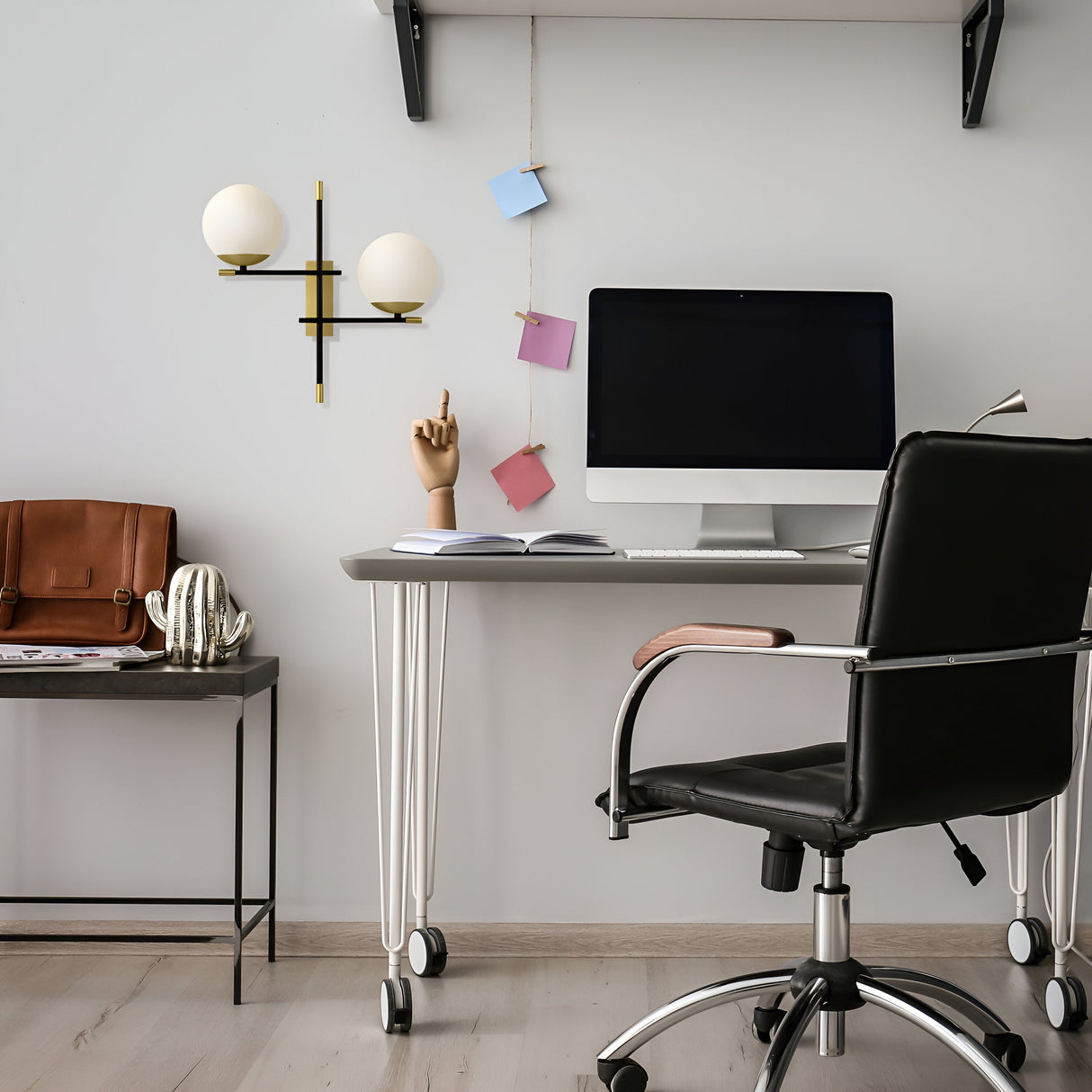 A modern workspace includes a desk with a computer, a sleek black office chair, and the Nostalgia 2 Light Wall Light - Black & Gold (MOD048WL-02G) for charm. Notable items are decorative hand sculptures, blue and pink sticky notes on the wall, a brown leather bag on a side table, and black and gold accents.