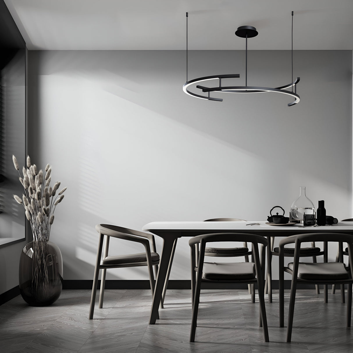A modern dining room with a minimalist design includes a sleek table, four chairs, and tall dried plants in a vase. The Line LED Circular Suspension 3000K - Black light, adjustable in height, illuminates the space with muted tones and natural light from a nearby window.
