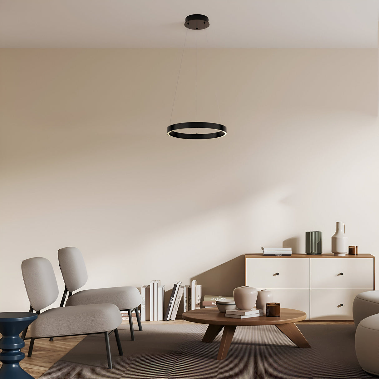 A minimalist living room with two beige chairs, a round wooden coffee table, and a cabinet holding books and decor. A Rim LED Circular Suspension Light 40cm 3000K - Black hangs from the ceiling, while light beige walls and a rug enhance the rooms cozy ambiance.