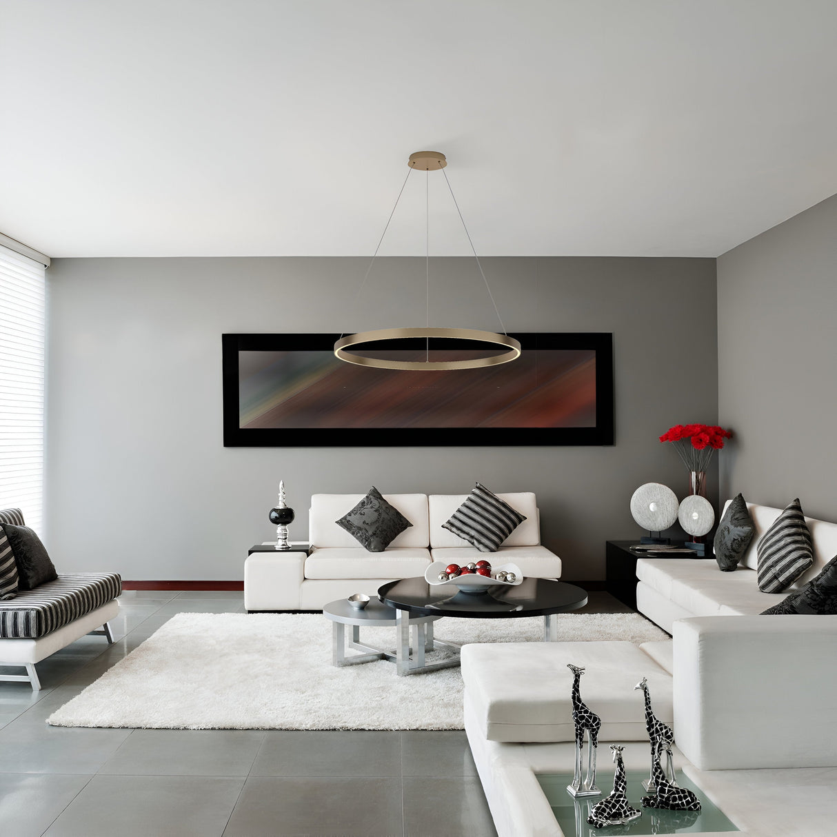 A modern living room features gray walls, a white sectional sofa, and zebra-patterned chairs. A round coffee table rests on a white rug. Abstract art enhances the space, while the Rim LED Circular Suspension Light 80cm 3000K in brass provides a warm glow. Vases and giraffe figurines add decorative touches.