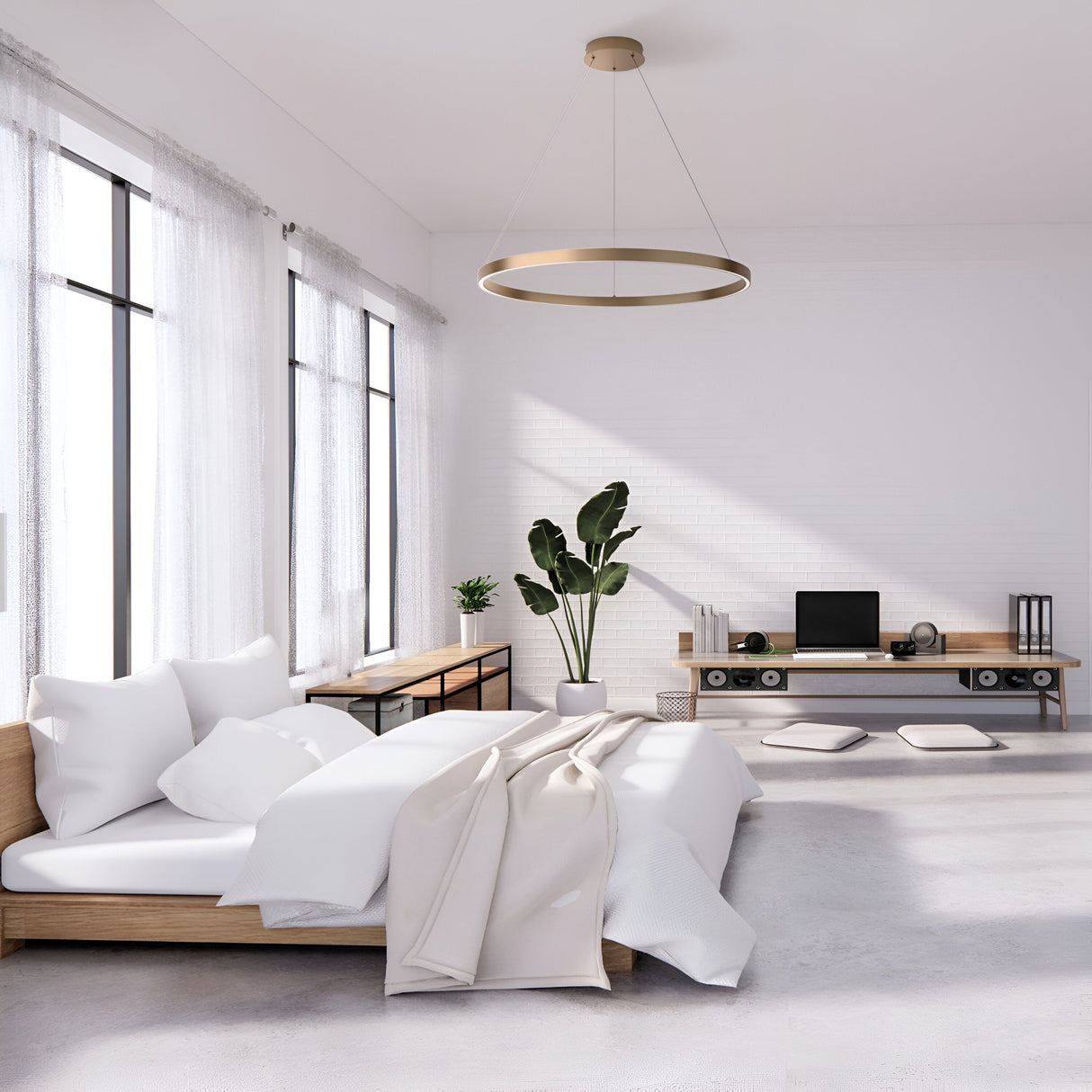 A minimalist bedroom features a wooden bed with white bedding and a beige throw, a plant beside it. On the right is a desk with a computer and speakers. The Rim LED Circular Suspension Light 80cm 4000K - Brass hangs overhead, while large windows fill the room with natural light.