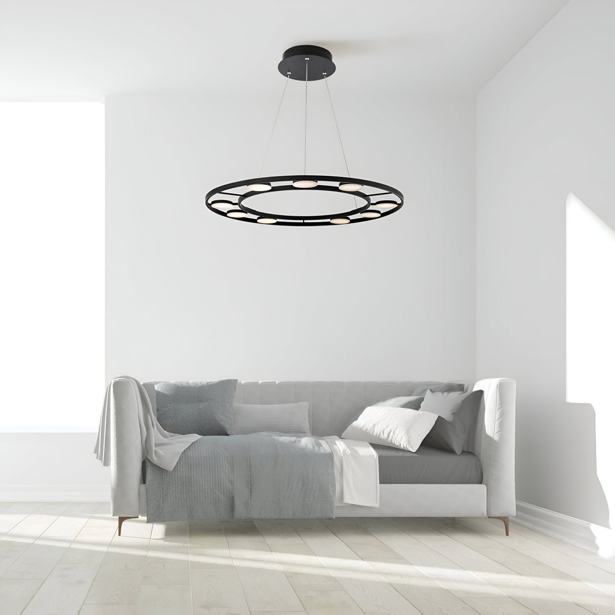 A minimalist living room showcases a modern touch with the Fad LED Circular Suspension Light in black hanging from the ceiling. Below is a white sofa with gray and white cushions and a draped blanket. Sunlight enters through a large window on the left, highlighting the light wood floor.