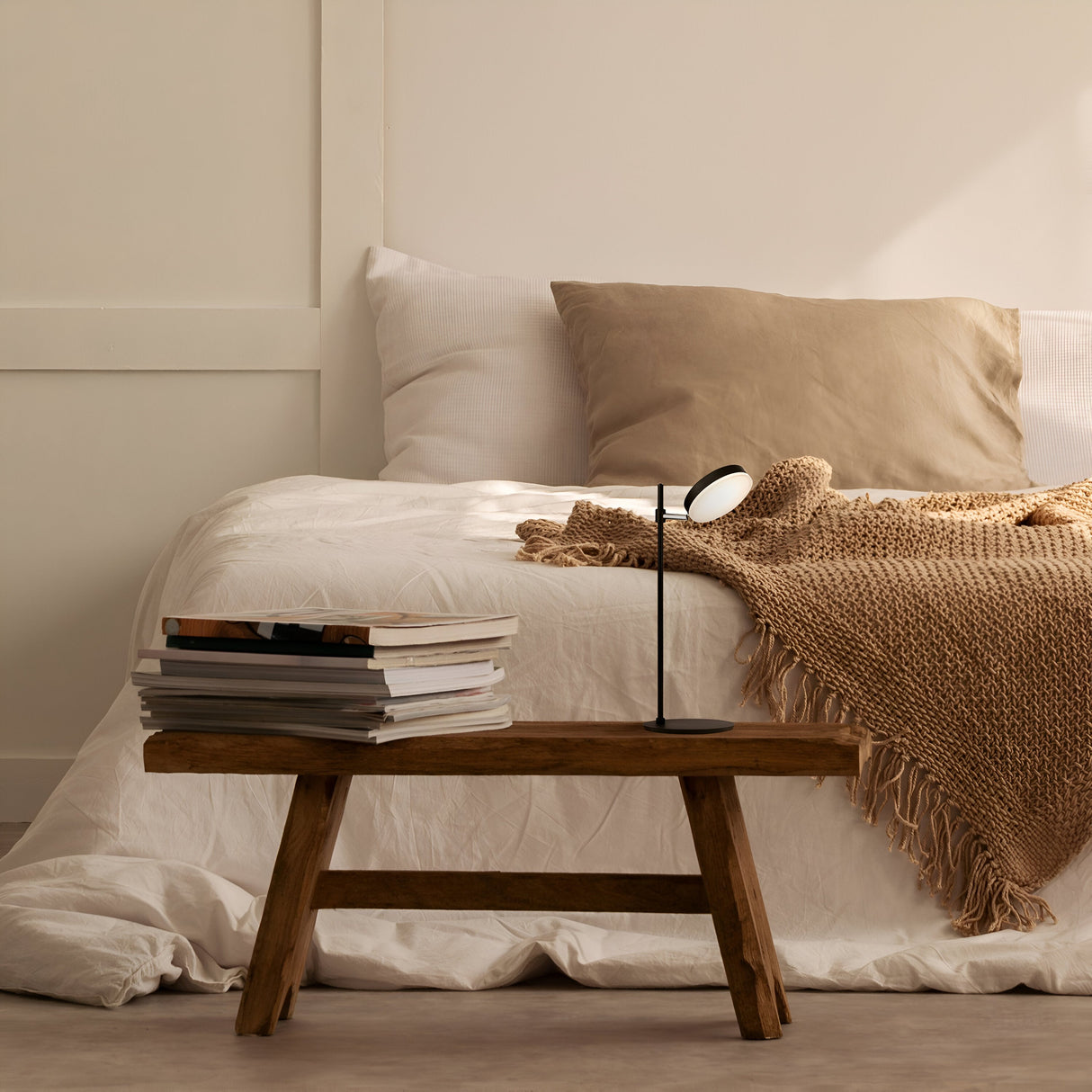 A cozy bedroom features a neatly made bed with white linens and a beige throw. A wooden bench beside the bed holds a stack of books and the Fad LED Table Lamp - Black. The rooms warm, neutral tones create a serene atmosphere, with energy-efficient lighting enriching the ambiance.