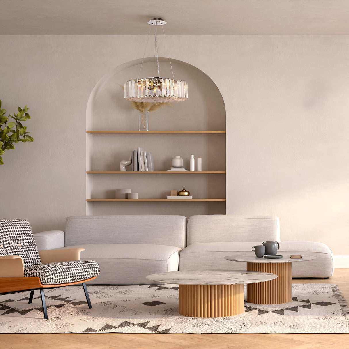 A modern living room features a white sofa, patterned armchair, and round wooden coffee tables on a geometric rug. Decor adorns built-in shelves in an arched alcove. The Recinto 6 Light Chandelier - Chrome illuminates the space, while a small plant adds greenery.