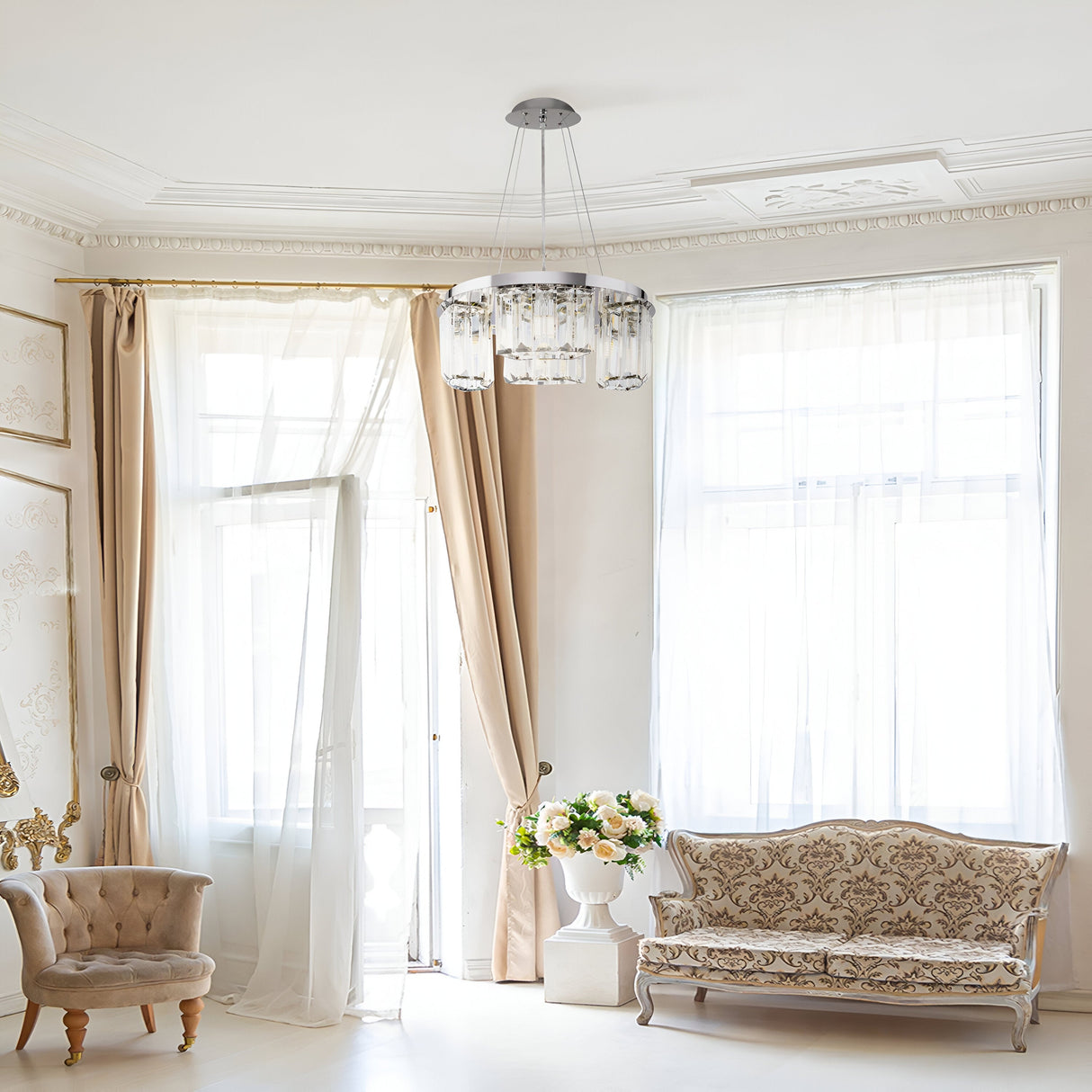 A bright, elegant room showcases large windows with sheer curtains, a Colline 4 Light Chandelier in chrome, a beige armchair, an ornate sofa, and a floral arrangement on a small table. The walls and ceiling feature decorative moldings.