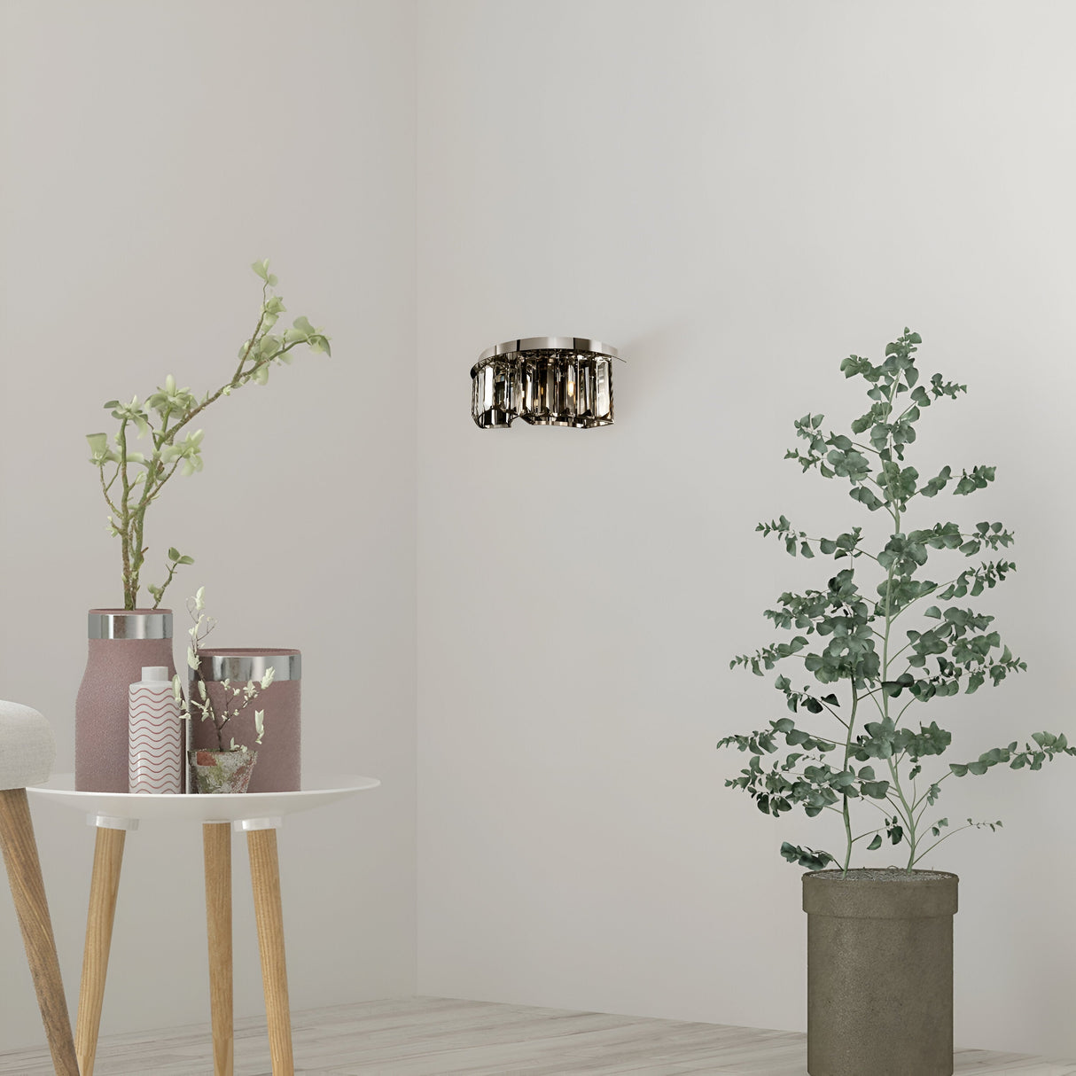In a minimalist room, the Colline Wall Light - Chrome accents a potted eucalyptus and a small round table. The table displays two pink and silver jars and a white-flowered plant. The light wood floor complements the stark white walls.