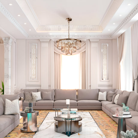 Elegant living room with a large U-shaped gray sectional sofa, ornate white walls, and a decorative ceiling. A Revero 7 Light Chandelier in gold hangs overhead, while a marble coffee table sits on a patterned rug. Natural light streams through tall windows.