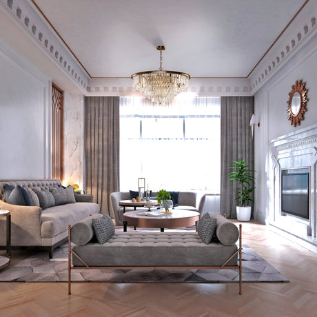 A luxurious living room features the Revero 16 Light Brass Chandelier with crystal pendants, a cozy gray sectional sofa, tufted bench, round coffee table, and large windows with curtains. A decorative fireplace is on the right and a potted plant adds greenery.