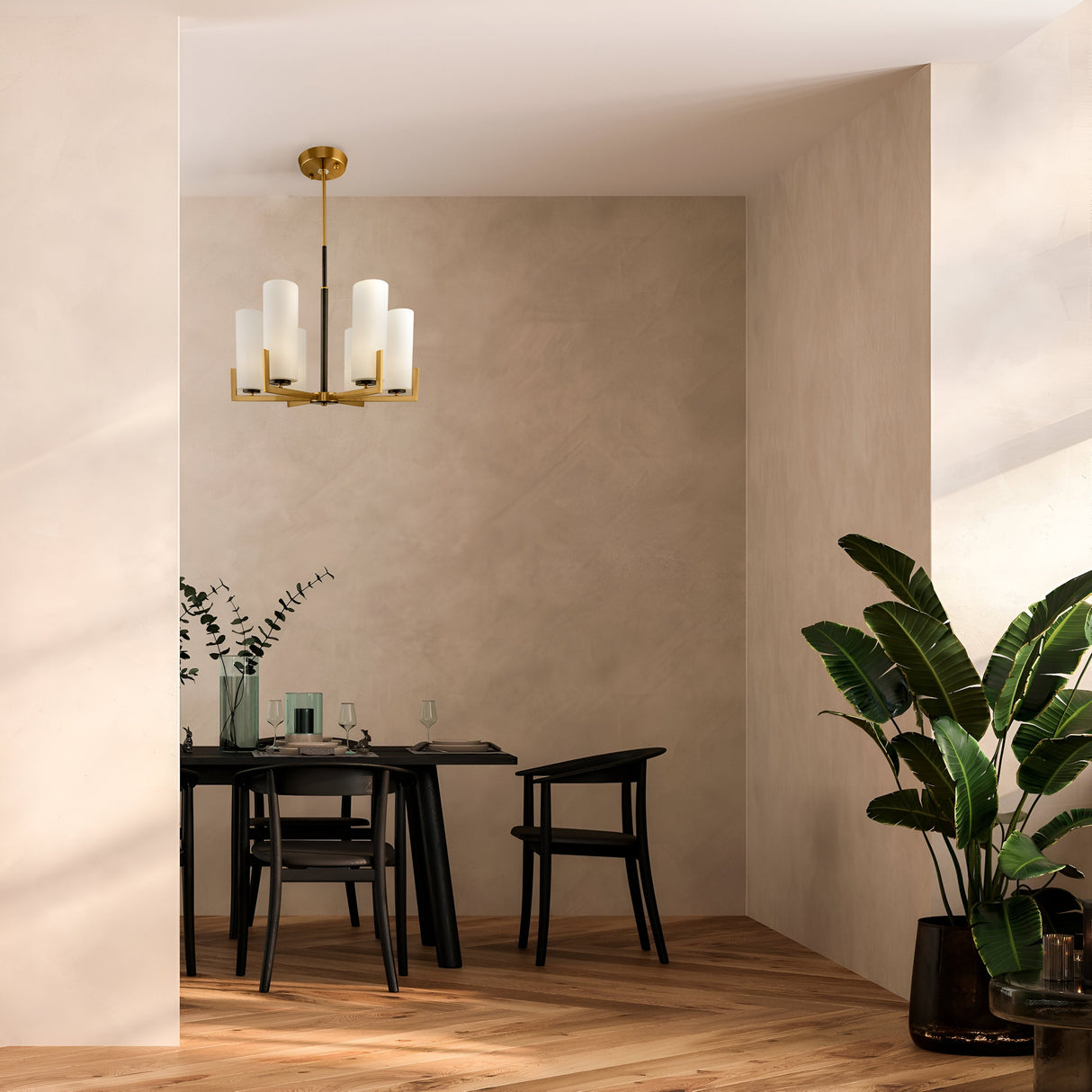 A minimalist dining space has a sleek black table and chairs against a beige wall. Above, the Fortano 6 Light Chandelier in brass adds a modern industrial touch. A black vase with green foliage adorns the table, while a large potted plant with broad leaves sits on the wooden floor.