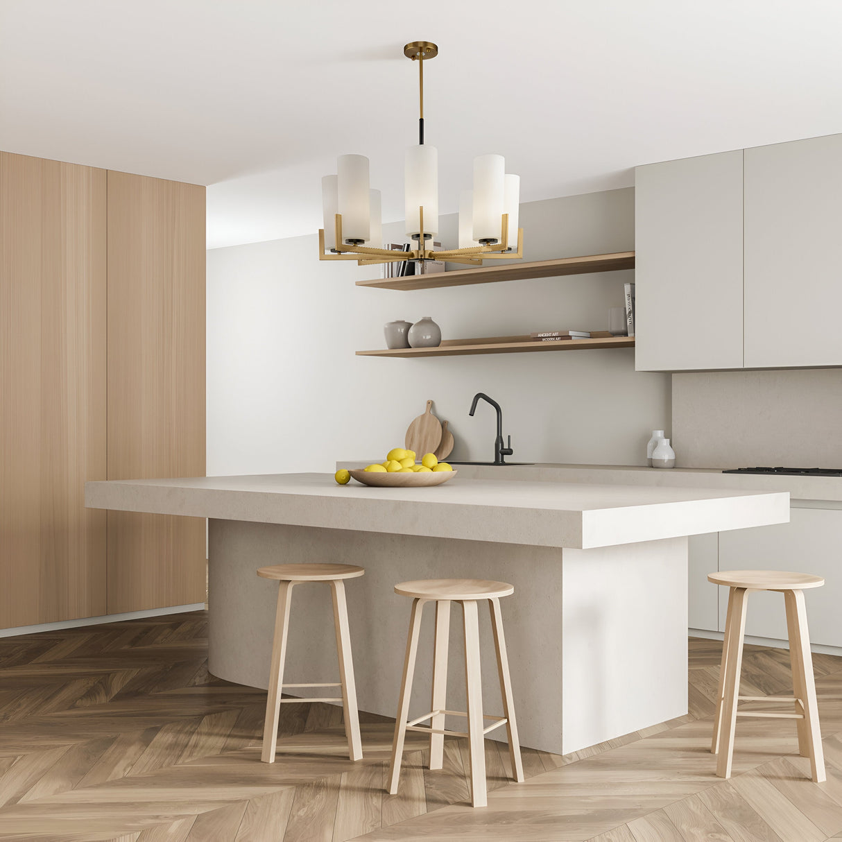 A modern kitchen features a large white island with three wooden stools, sleek wall cabinets, minimalist shelves for decor, and a black faucet. The Fortano 8 Light Chandelier in brass adds style above the island where a bowl of lemons sits.