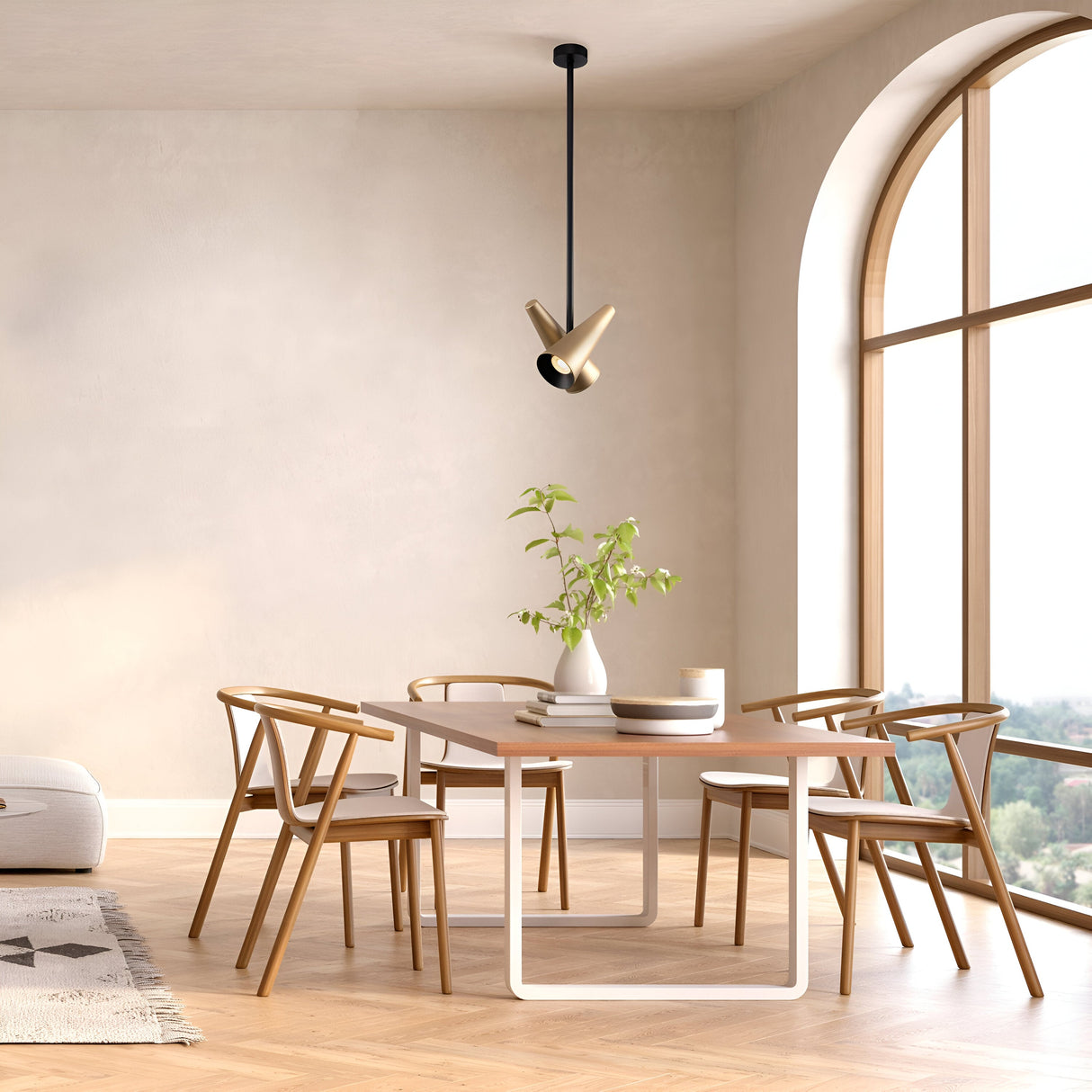 A modern dining room showcases a light wooden table, four matching chairs, and a white vase with greenery. The Giro 2 Light Adjustable Heads Pendant in brass adds an industrial touch. Large arched windows on the right flood natural light onto the chevron-patterned floor.