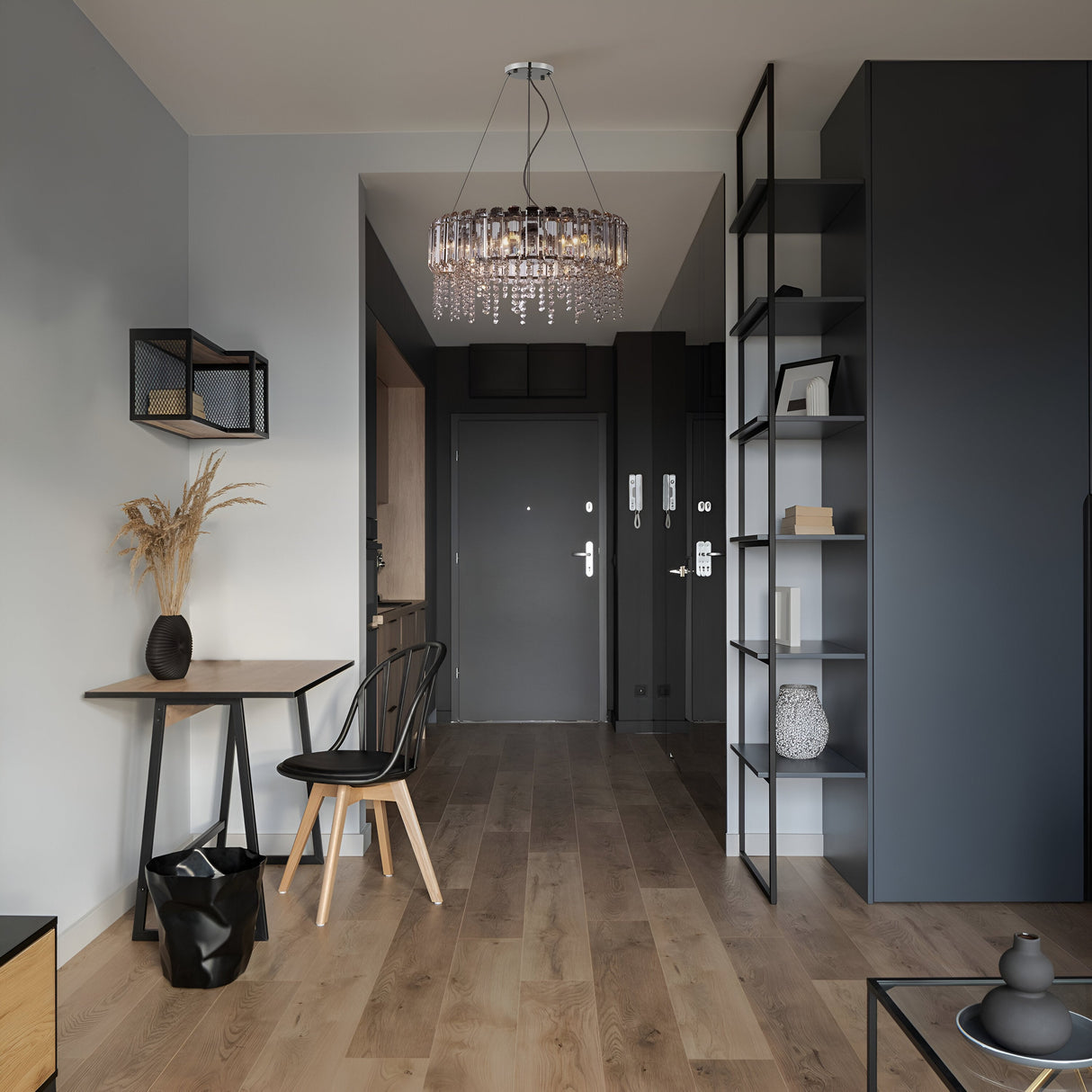 A modern hallway features dark hardwood floors, gray walls, and a black door. Complemented by open shelving, a small table, chair, and black vase with dried plants. Tying it all together is the Hail 8 Light Chandelier - Chrome, sparkling with crystal accents in the minimalist decor.