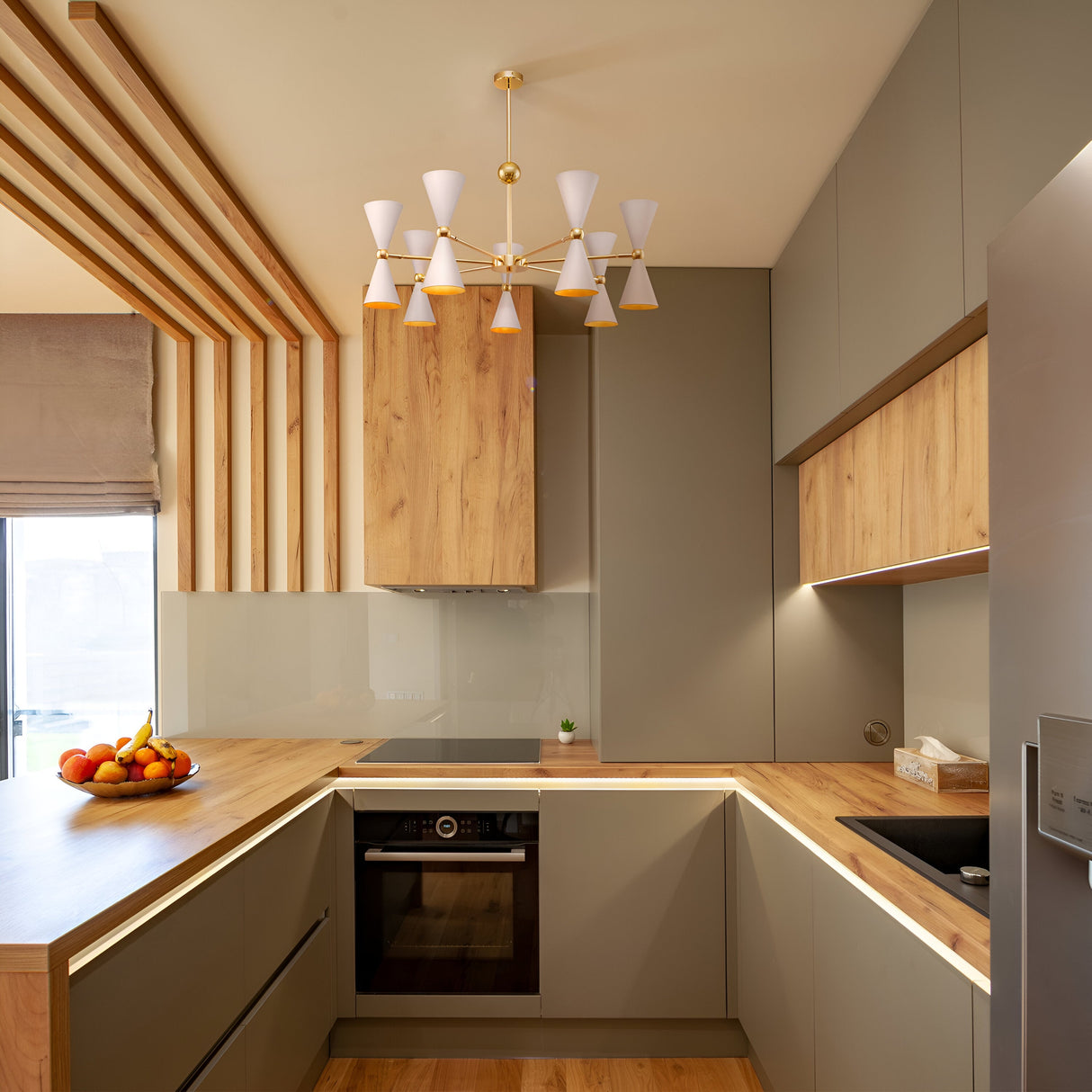 This modern kitchen features light wood cabinets, gray cupboards, and a built-in oven. A fruit bowl sits on the counter. The minimalist design is highlighted by the Vesper 14 Chandelier - Gold & White, adding elegance with its multiple cone-shaped lights.