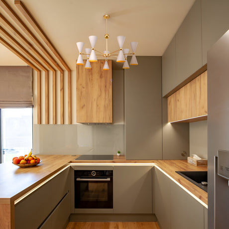 This modern kitchen features light wood cabinets, gray cupboards, and a built-in oven. A fruit bowl sits on the counter. The minimalist design is highlighted by the Vesper 14 Chandelier - Gold & White, adding elegance with its multiple cone-shaped lights.