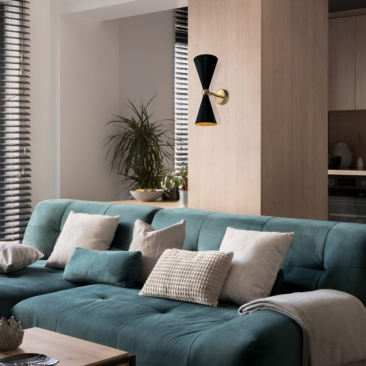 A modern living room showcases a green velvet sofa with beige and cream pillows. A potted plant rests on a light wood cabinet behind it. Gold & Black window blinds filter sunlight, while a decorative Vesper Up & Down Rotable Wall Light in gold and black completes the look.