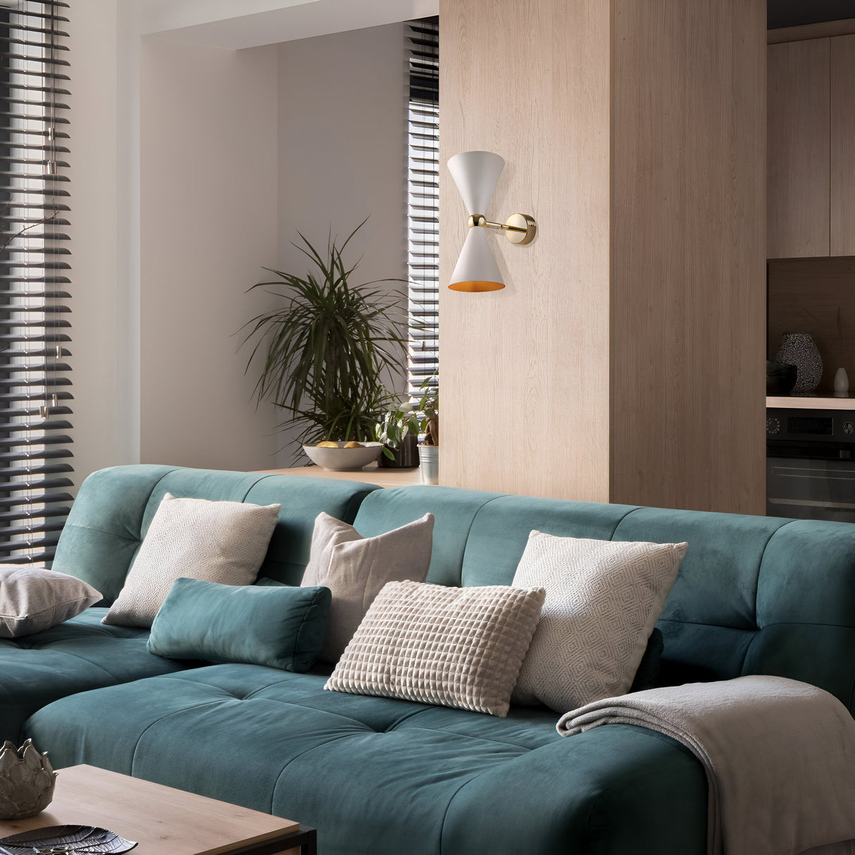 A cozy living room showcases a green sofa with white and beige pillows. Above, the Vesper Up & Down Rotable Wall Light in gold and white adds elegance. In the corner near blinds, a potted plant thrives, while a wooden divider separates the space artfully.