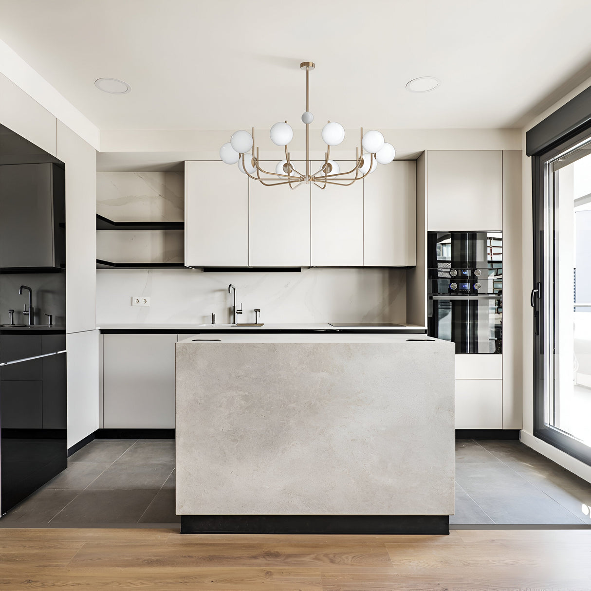 Modern kitchen with white cabinets, black accents, and a central island with a light gray countertop. A Rendez-vous 10 Light Chandelier in brass stylishly hangs above. The floor highlights dark and light wood patterns, with a large window on the right inviting natural light.