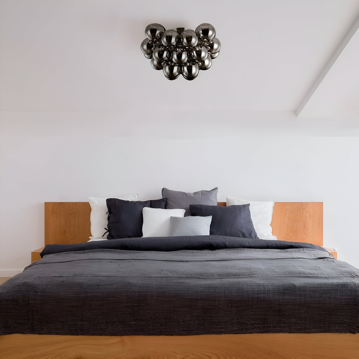 A modern luxury bedroom has a wooden bed frame with dark gray bedding and decorative pillows in gray and white. A unique Balbo 8 Light Semi-Flush chandelier in Nickel with smoky glass complements the minimalist design and clean white walls from its spot on the ceiling.