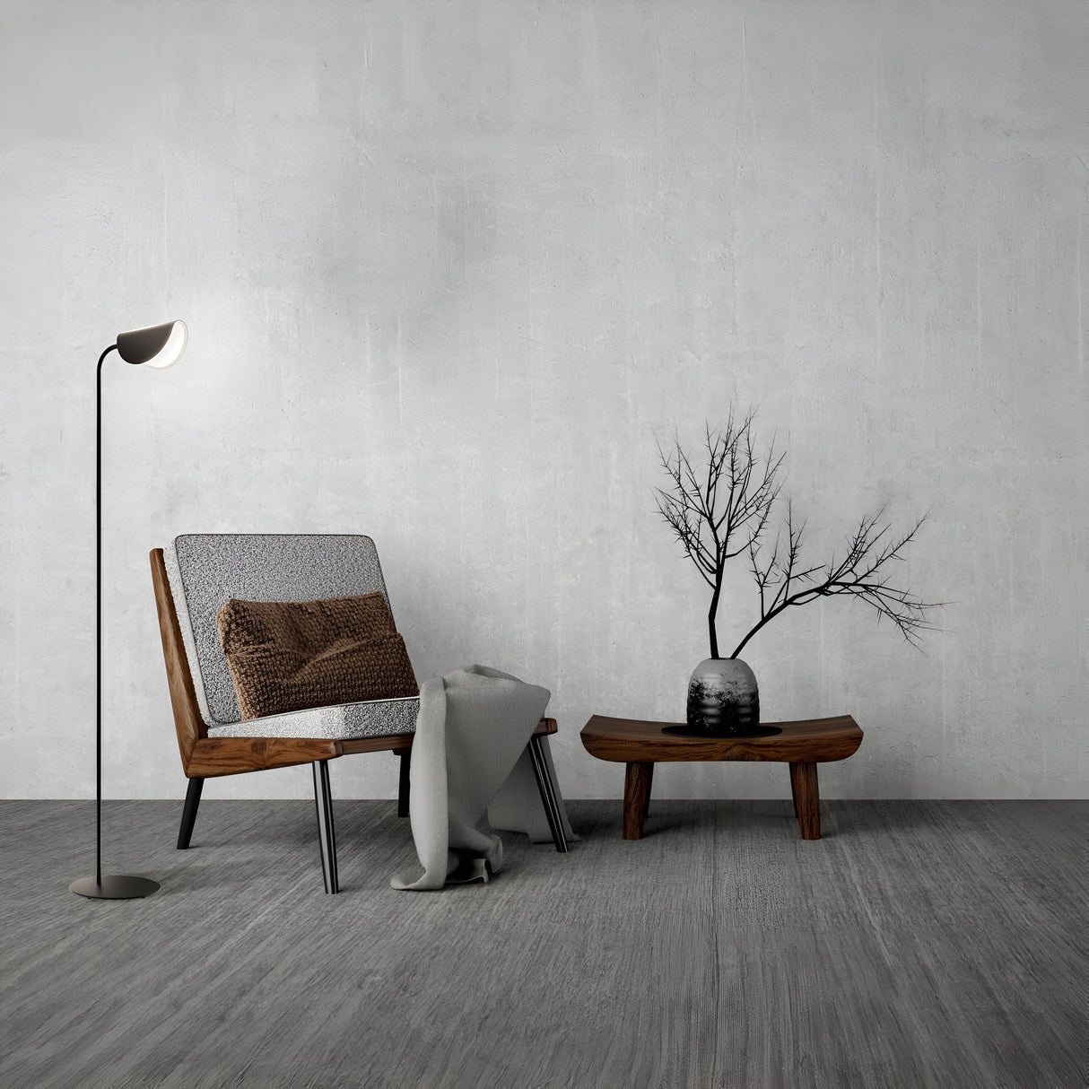 A minimalist living space features a wooden chair with a gray cushion, a cozy blanket, and the sleek Mollis Floor Lamp in black for ambiance. A small table holds a black vase with bare branches, complementing the modern interiors with gray walls and flooring.