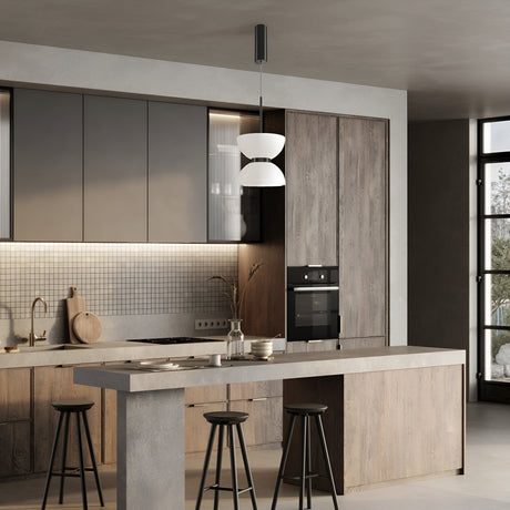 This modern kitchen features a concrete island, three black bar stools, and wooden cabinets. A built-in oven sits on the right, and the backsplash has small tiles. Kyoto LED Pendant Light - Black With Glossy White Glass provides energy-efficient lighting, complemented by natural light and dried plants decor.