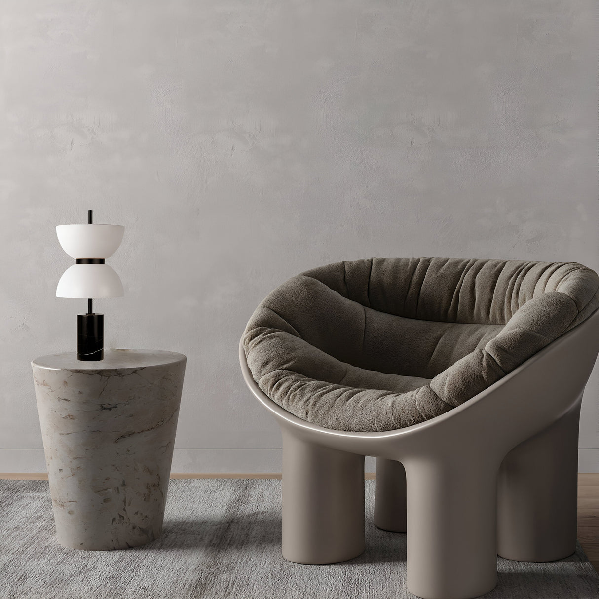 A modern plush gray chair with an arched design sits by a round marble side table, which supports the Kyoto LED Table Lamp - Black. This minimalist setup rests on a gray rug against a textured wall.