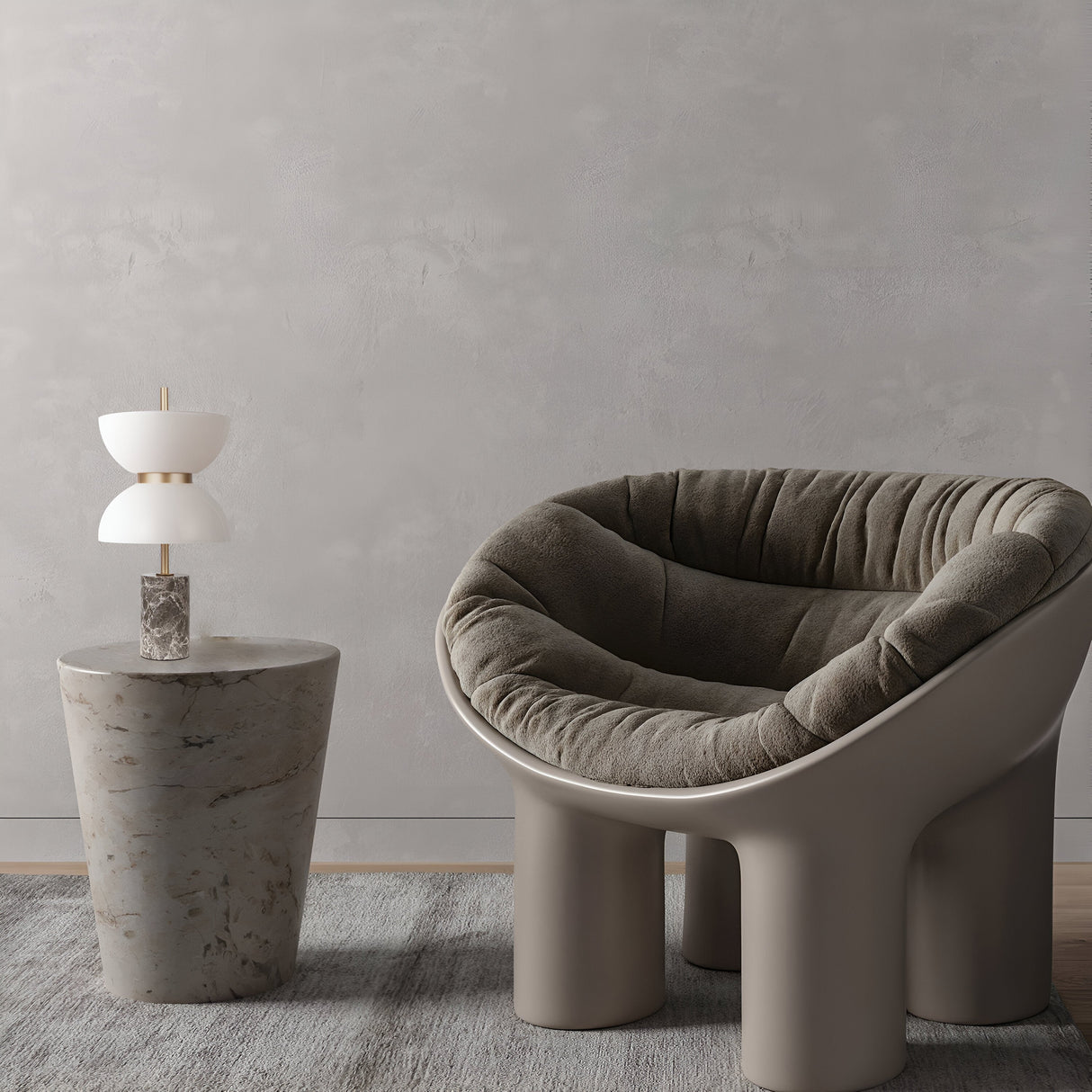 A modern, plush gray chair with a rounded shape sits on a textured gray rug. To its right, a small side table holds the Kyoto LED Table Lamp - Gold & Grey. The background features a textured gray wall, adding depth and elegance to the serene setting.