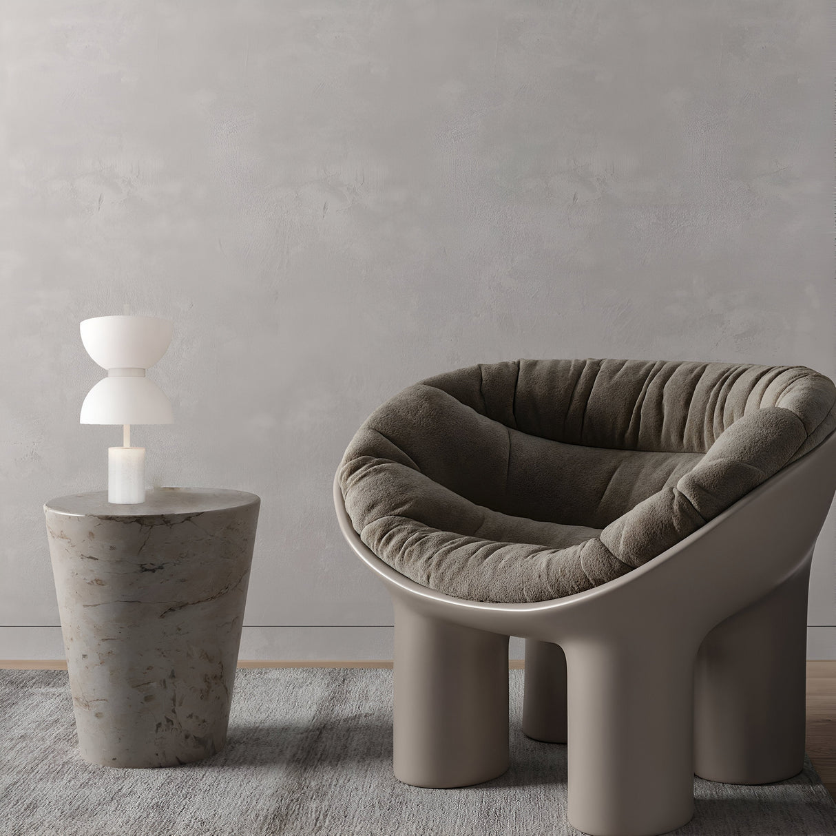A plush, curved armchair with textured fabric is next to a round, stone-textured side table topped with the Kyoto LED Table Lamp - White, featuring a geometric design and marble base. The stylish arrangement sits on a gray rug against a light gray wall.