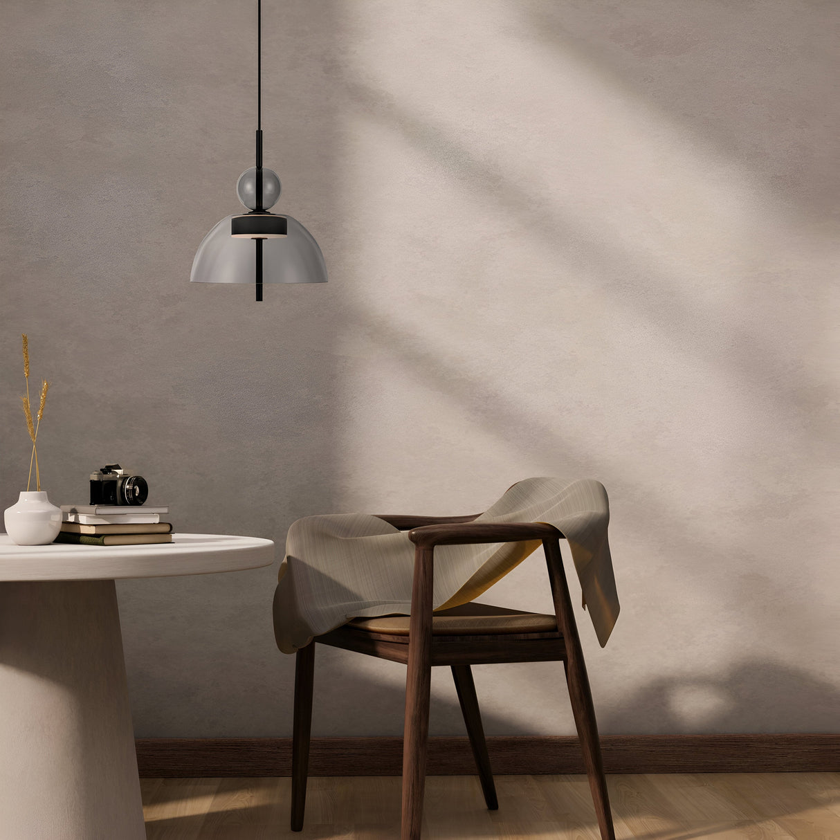 A minimalist room with a Bangkok LED Pendant Light 25cm - Black With Round Grey Glass Shade casting shadows on a textured wall. A round table holds a camera and vase, while a draped cloth rests on a wooden chair with curved backrest. Neutral tones of modern interiors create a calm ambiance.