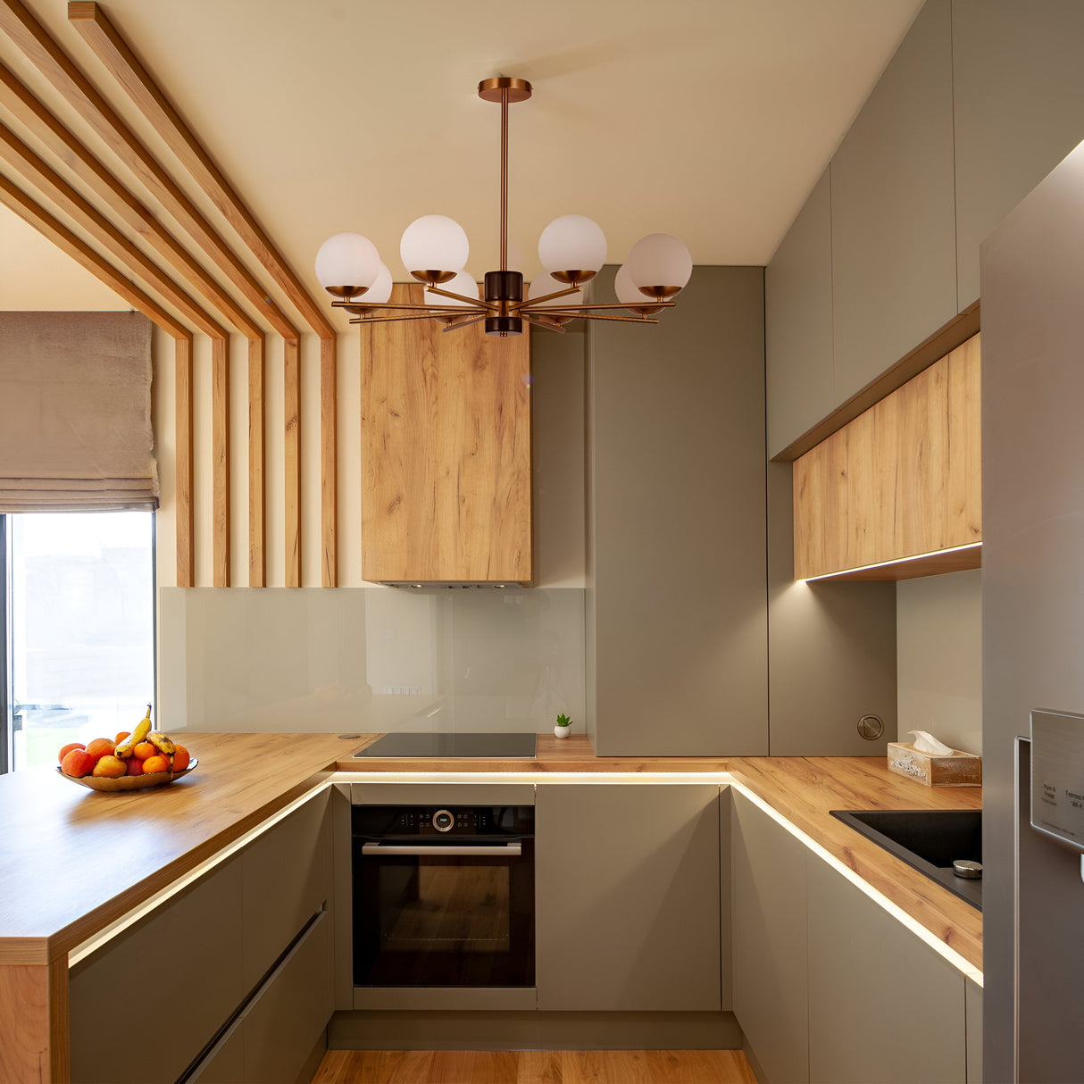 Contemporary kitchen features wooden countertops and cabinets, gray cabinetry, stainless steel appliances such as an oven and fridge, with a fruit bowl containing bananas and oranges on the counter. Overhead hangs the Marble 6 Light Chandelier in Brass with opal glass globes.