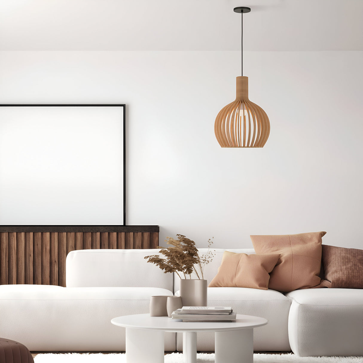A minimalist living room features a white sofa, beige and brown pillows, and a small white round table with dried flowers. Above hangs a Roots Pendant Light 35cm - Black with Beige Wooden Globe Dome Shade. A large blank picture frame decorates the wood-paneled wall, part of the Modern Nature collection.