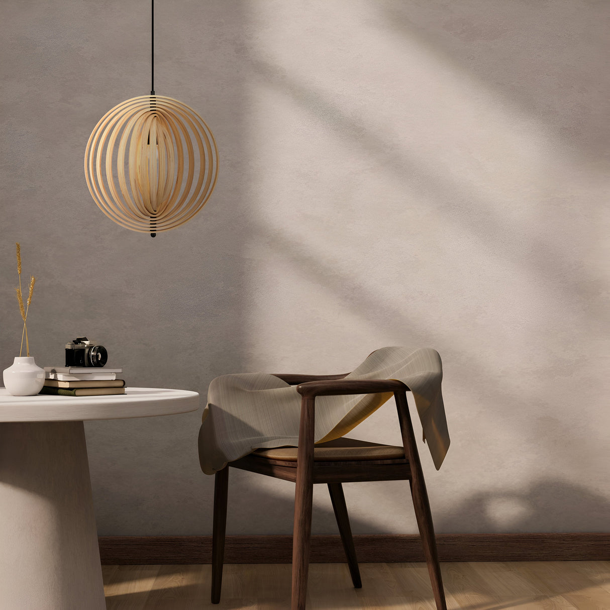 A minimalist room features a wooden chair with fabric, a round table holding books and a vintage camera, while the Roots Pendant Light 43cm - Black & Natural Wood Sphere Rings from the modern nature collection casts intricate shadows on the textured wall.