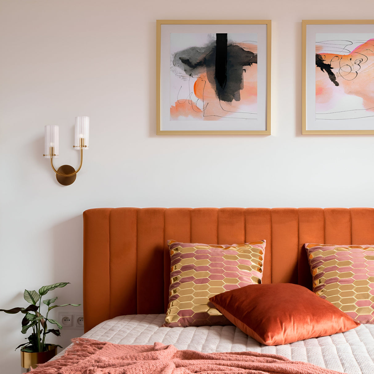 A bedroom features a burnt orange upholstered headboard with two geometric-patterned cushions and a rust pillow. Two abstract art pieces hang above the bed, while a small plant sits to the left beneath the Arco 2 Light Wall Light in brass with ribbed glass.