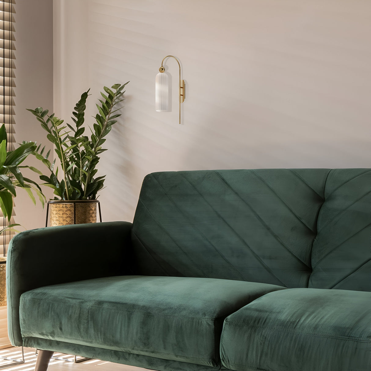 A cozy living room showcases a dark green velvet sofa with a nearby potted plant featuring broad leaves. The Antic Wall Light in gold with a ribbed white glass lampshade softly glows above, while wooden blinds on the window allow soft light to filter through.