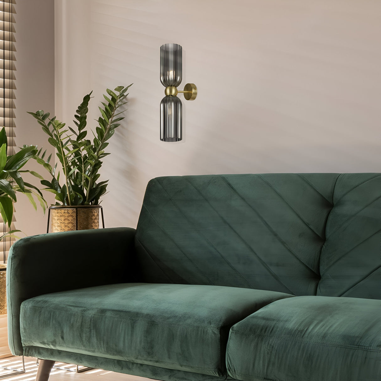 A modern living room showcases a green velvet sofa and the Antic 2 Light Wall Light, featuring gold accents and grey glass shades. Two potted plants are on the left, while sunlight filters through blinds, casting shadows on the cream-colored wall, echoing retro lighting trends.