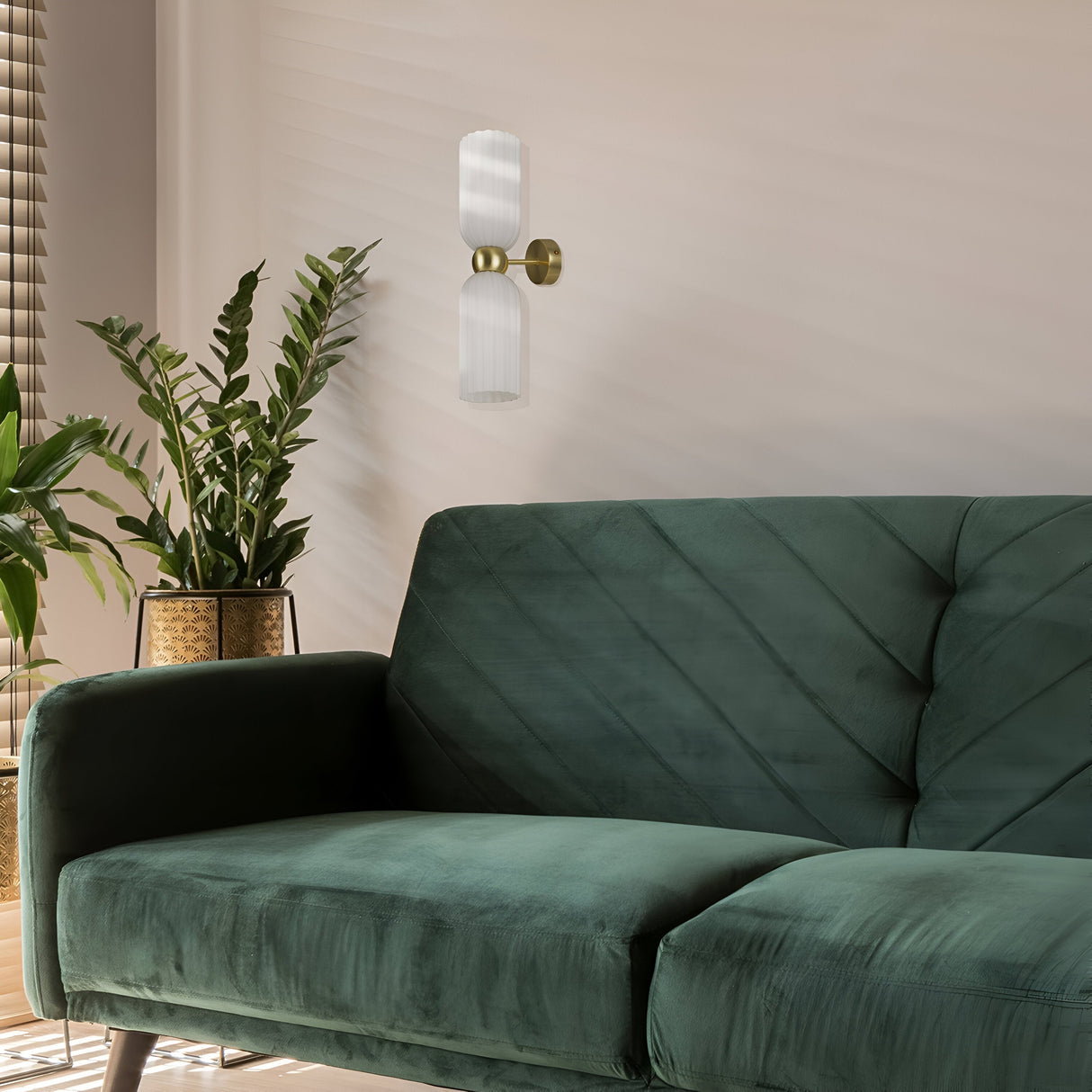 A modern living room showcases a deep green velvet sofa against a light wall, illuminated by the Antic 2 Light Wall Light in gold with white glass lampshades. A potted plant sits beside the sofa on the wooden floor, and blinds partially cover a sunlit window.