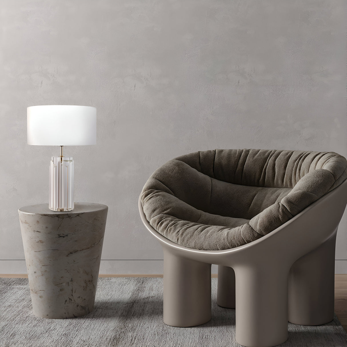 A modern living space features a plush, rounded gray armchair with a unique base and a cylindrical side table. The Muse Table Lamp - Gold With White Shade sits elegantly on the table, complementing the smooth gray wall and light-colored rug in this stylish design.