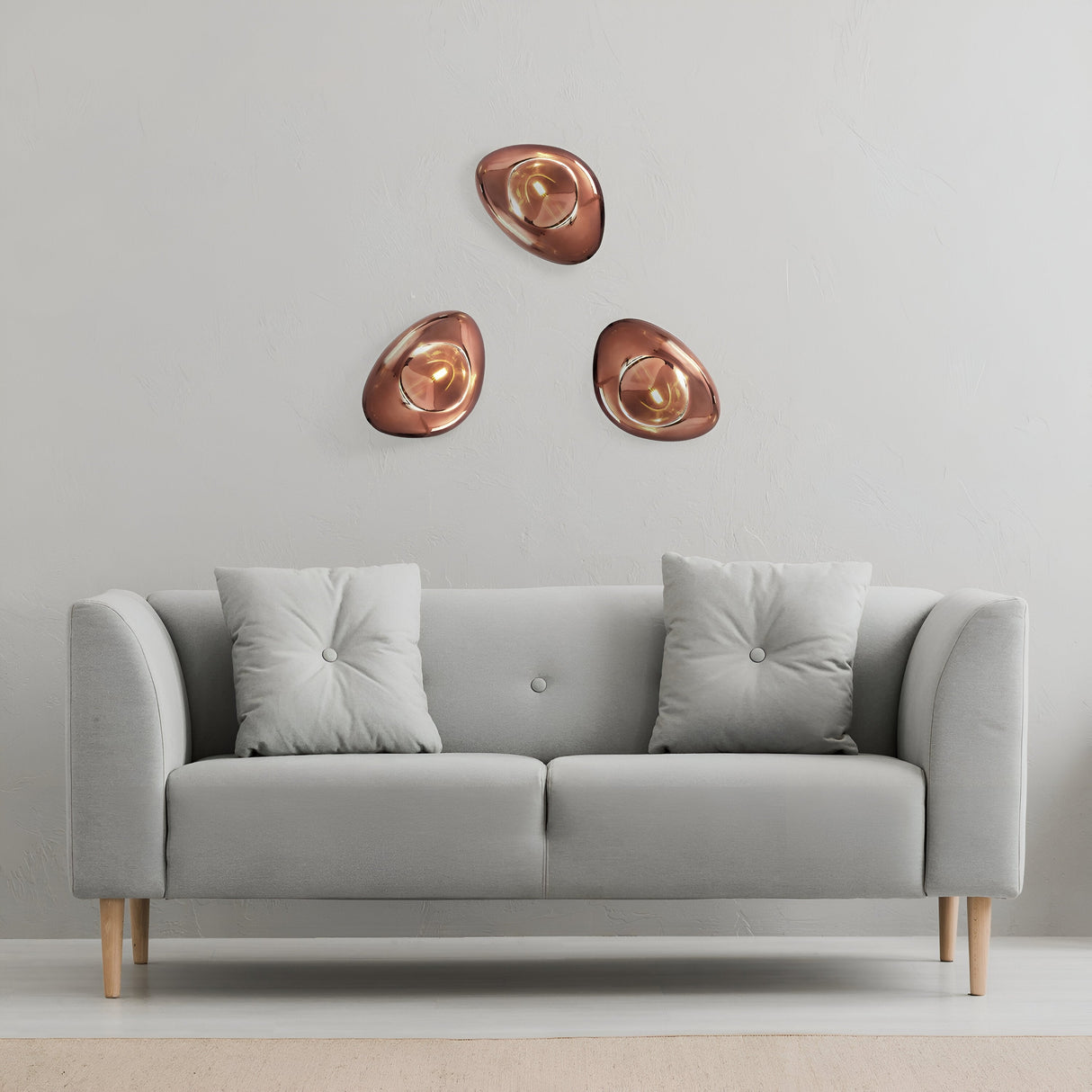 A minimalist room features a light gray sofa with two tufted cushions. Above it, Mabell Wall Lights in copper illuminate three abstract metallic wall sculptures, arranged in a triangular pattern on a light-colored wall.