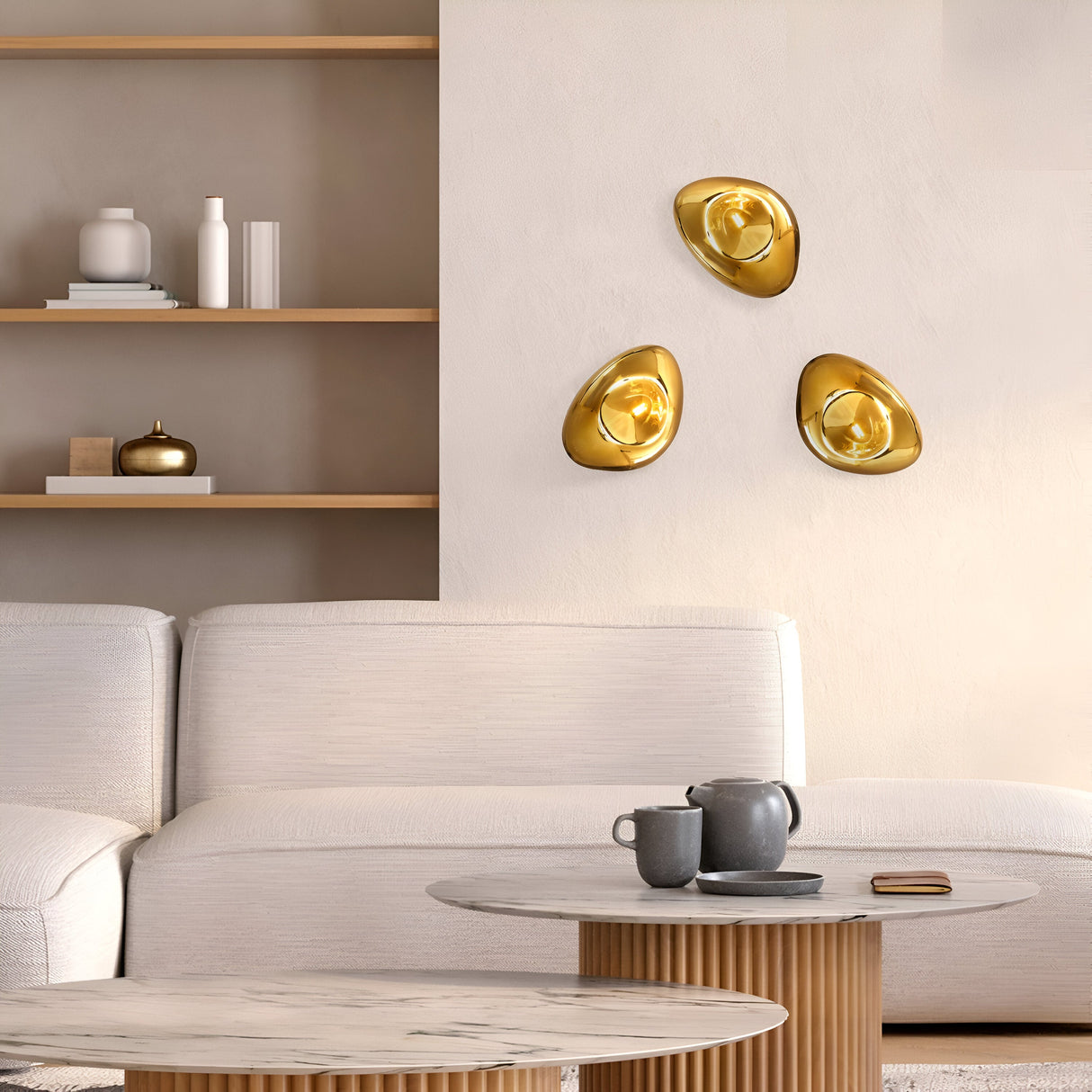 A modern living room showcases a white sofa and wooden shelves with decor, enhanced by three gold abstract wall art pieces. A Mabell Wall Light - Gold offers ambient lighting, while a round marble-top coffee table supports a gray teapot and cup within the neutral color scheme.