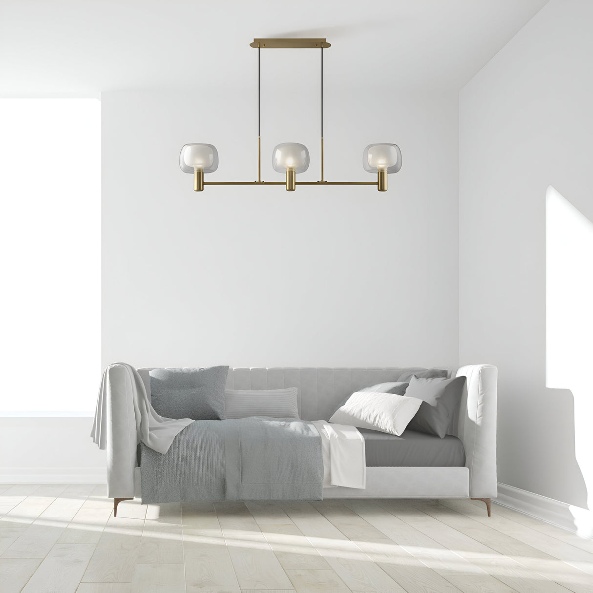 A minimalist living room showcases a white tufted couch with gray and white pillows and a throw. The Vision 3 Light Suspension in gold, featuring inner frosted and outer clear glass, elegantly hangs above. Sunlight streams through a large window, casting shadows on the hardwood floor.