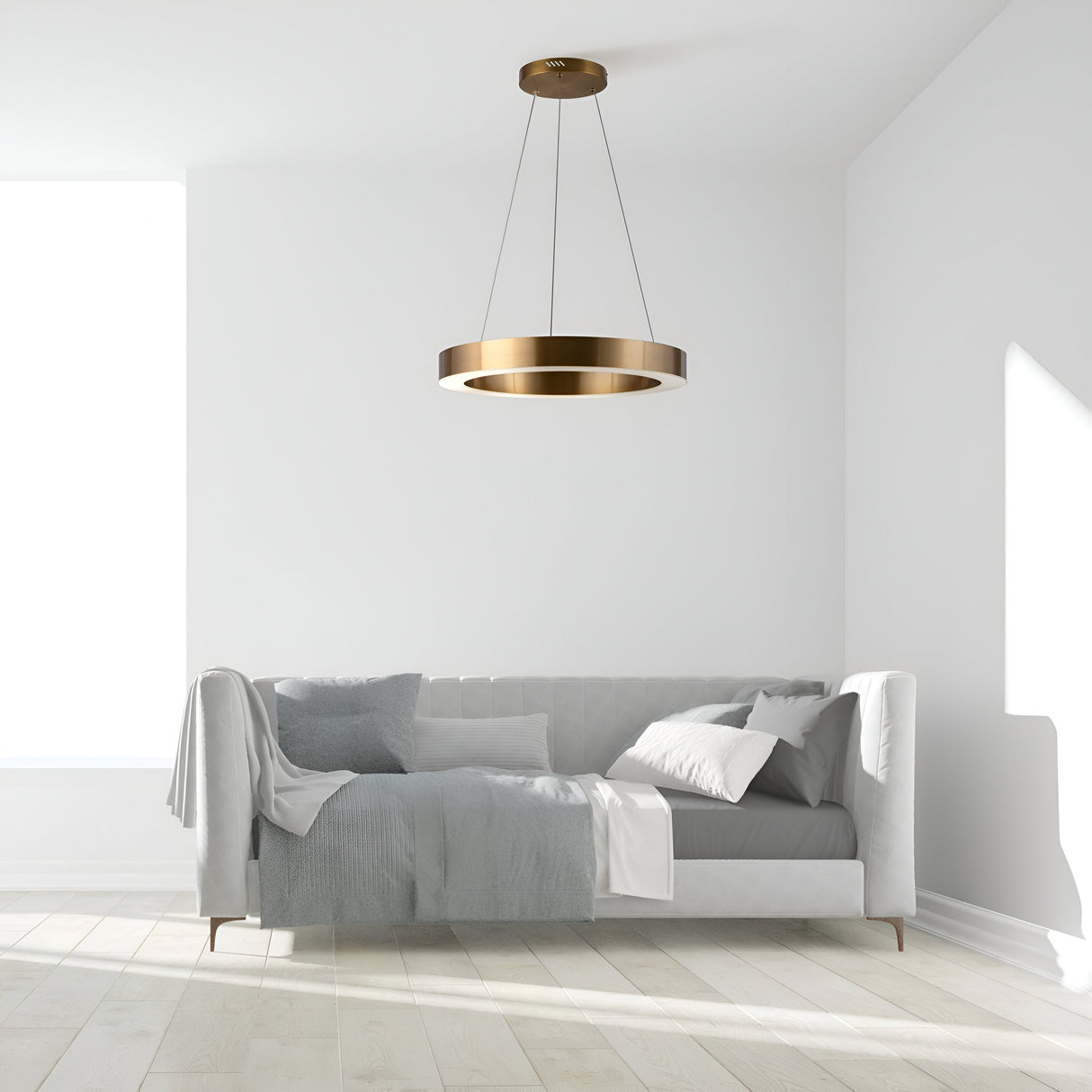 A minimalist living room boasts a white sofa with gray and white pillows and a throw blanket. The Saturno LED Circular Suspension Light 40cm - Brass, featuring energy-efficient LED technology, hangs from the ceiling. Light wood floors and a bright window complete the airy vibe.