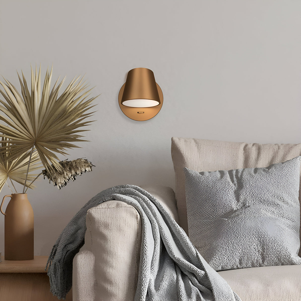 A cozy living room has a light gray sofa with a textured throw and pillow. A brown vase with dried leaves sits beside it, while the Pixel LED Wall Light - Brass decorates the plain wall above.