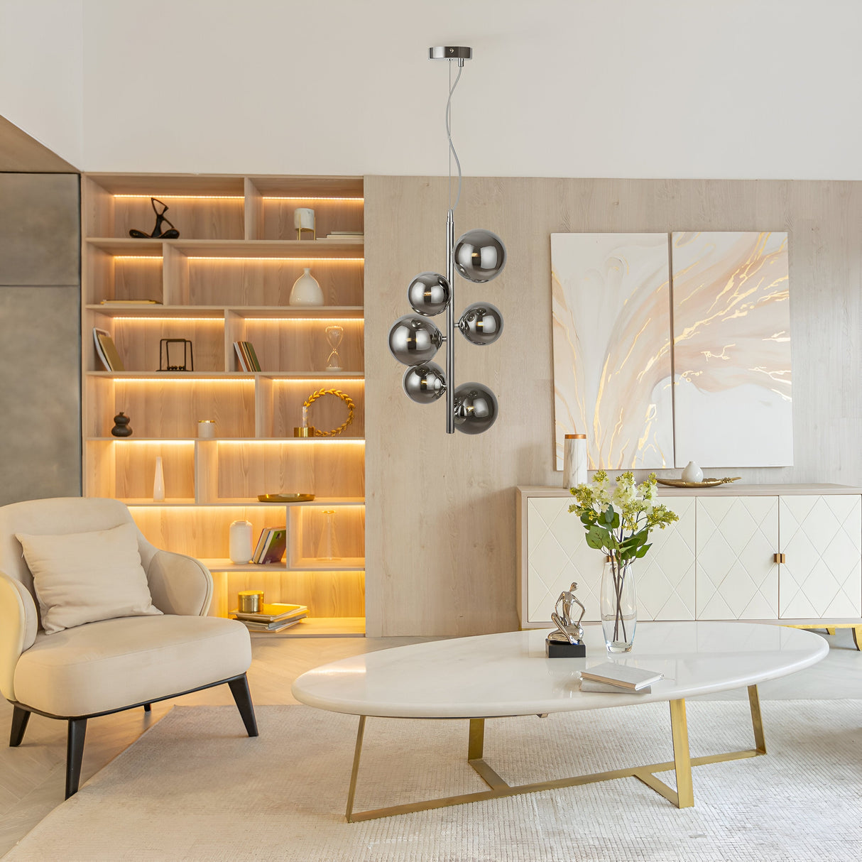 A modern living space features a beige armchair, white oval coffee table, abstract wall art, and the Dallas 6 Light Drop Suspension with smoked mirrored glass spheres overhead. Warmly lit bookshelves and a light-colored sideboard complete the elegant decor.