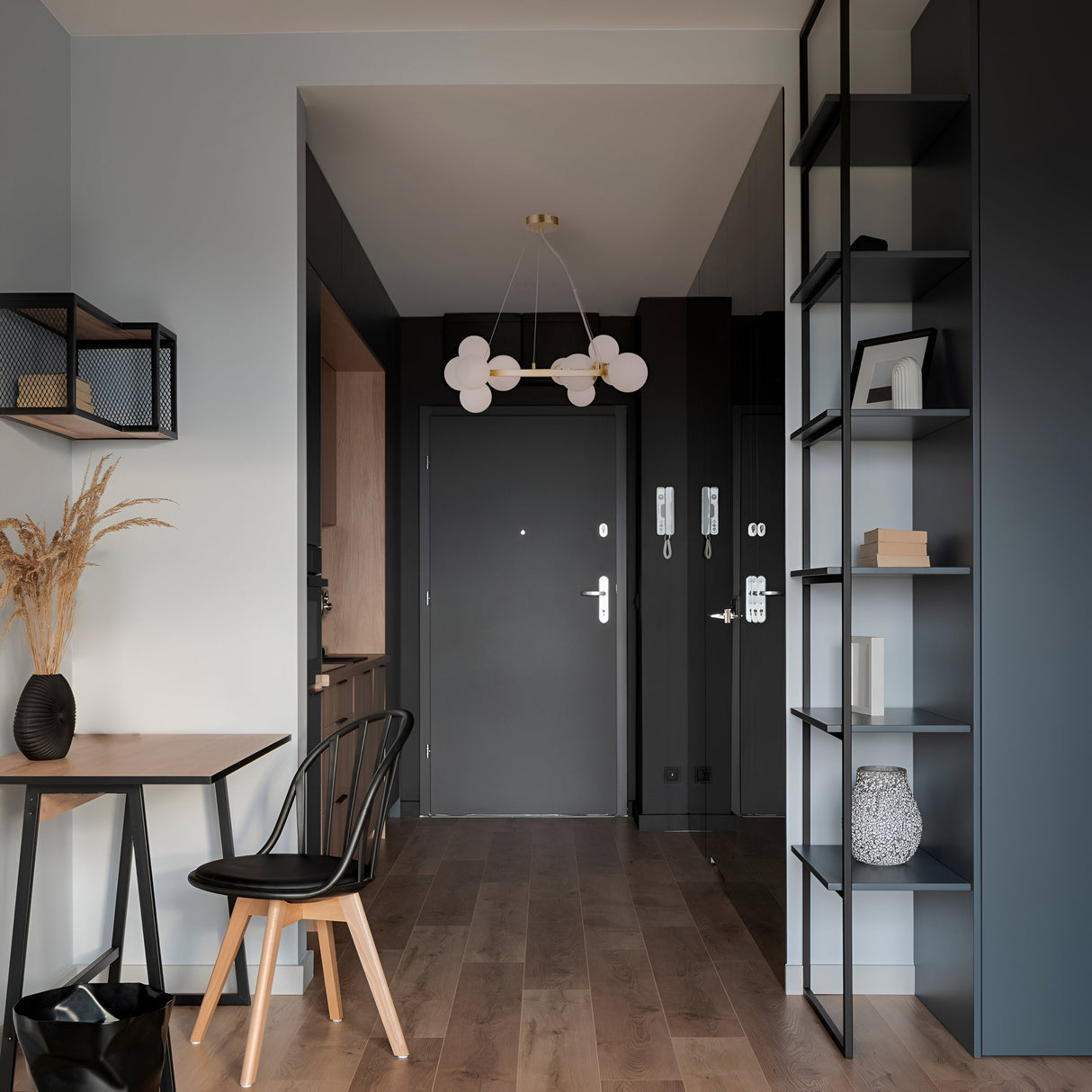 A stylish entryway boasts a dark door, bubble chandelier with adjustable cables, and wooden floor. A black chair and table with decor are on the left, while a tall shelving unit displays items on the right. The Dallas 11 Light Suspension in gold with white glass enhances the minimalist charm.
