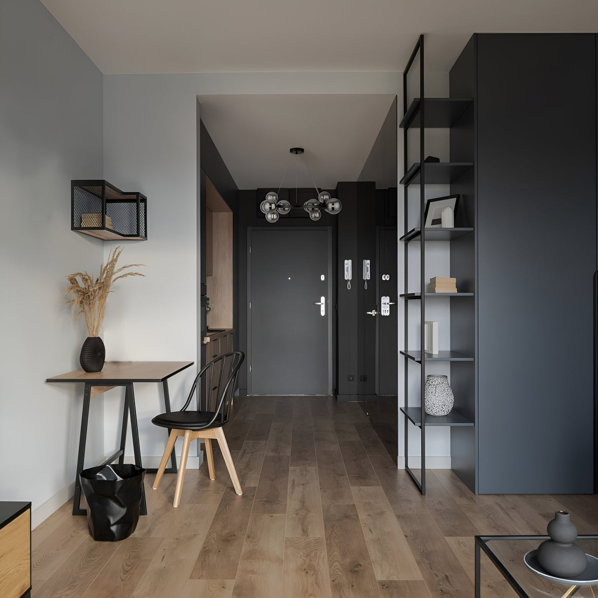 A minimalist modern interior features a small wooden table with a black chair, a vase containing dried plants, and a metal shelf. The Dallas 11 Light Suspension in Black with Smoked Glass illuminates light and dark gray walls, complementing the wooden floor and contemporary aesthetic. A door is visible in the background.