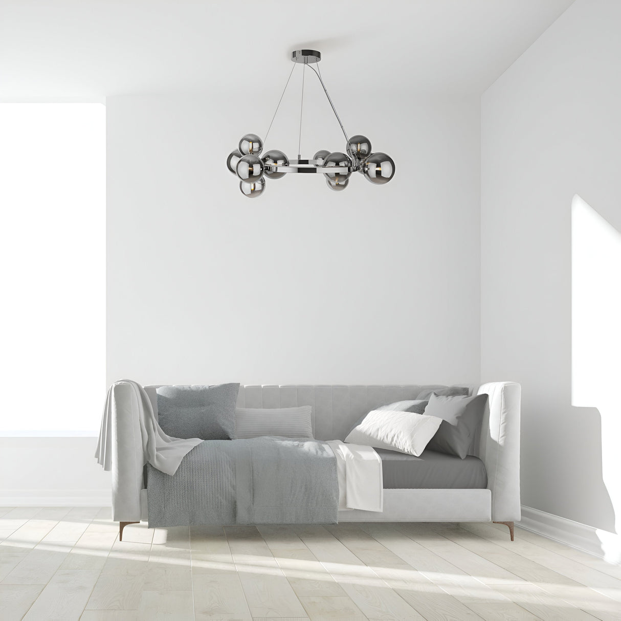 A minimalist living room features a modern gray couch adorned with assorted pillows and a throw blanket. The Dallas 11 Light Suspension in chrome with smoked glass, featuring spherical bulbs, hangs above. Natural light floods through a large window onto the light wood floor.