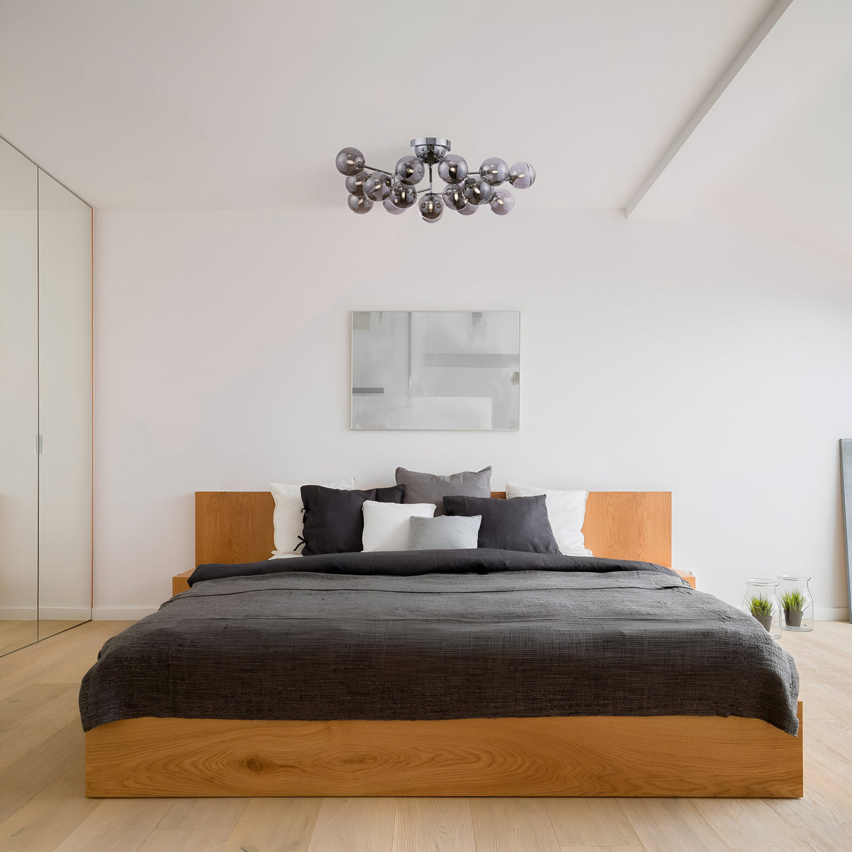 A minimalist bedroom includes a wooden bed frame, dark gray bedding, white and gray pillows, and light wooden flooring. The Dallas 20 Light Semi-Flush Chrome with Smoky Glass chandelier is suspended above. A glass wall lines one side of the room, with abstract art on the wall behind the bed.