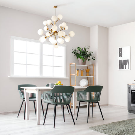A contemporary dining space includes a white table and four dark green chairs, enhanced by the elegant Dallas 24 Light Chandelier in gold with white glass. A shelving unit with plants and a Be Happy sign adds charm, while abundant natural light flows through the window.