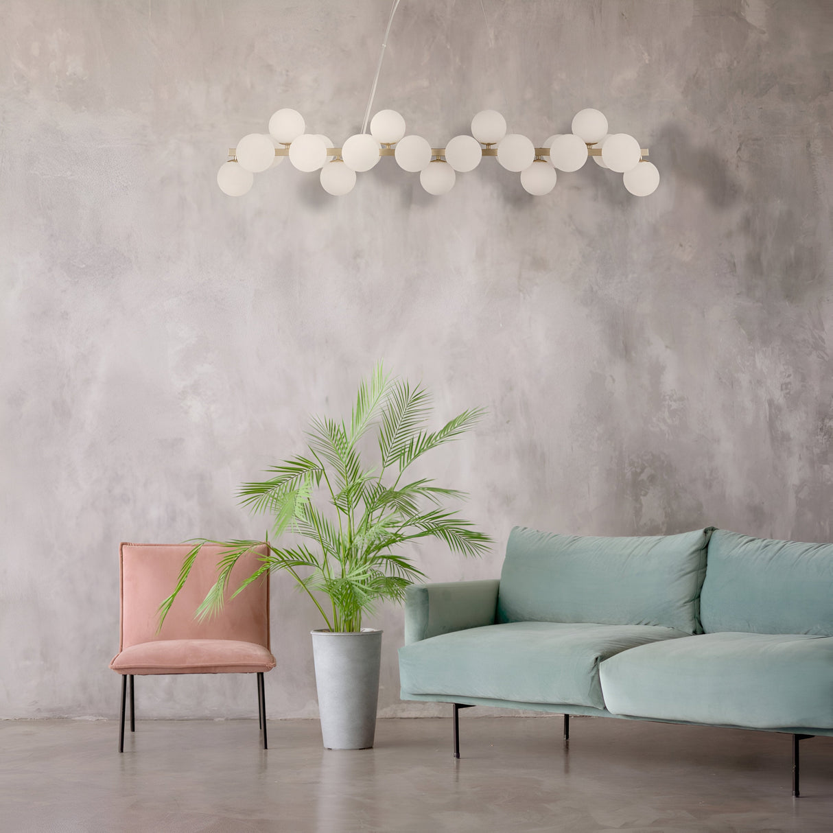 A modern living room has a gray textured wall, mint green sofa, pink velvet chair, and potted palm plant. The Dallas 25 Light Linear Suspension in gold with white glass spheres elegantly hangs from the ceiling, adding contemporary charm.