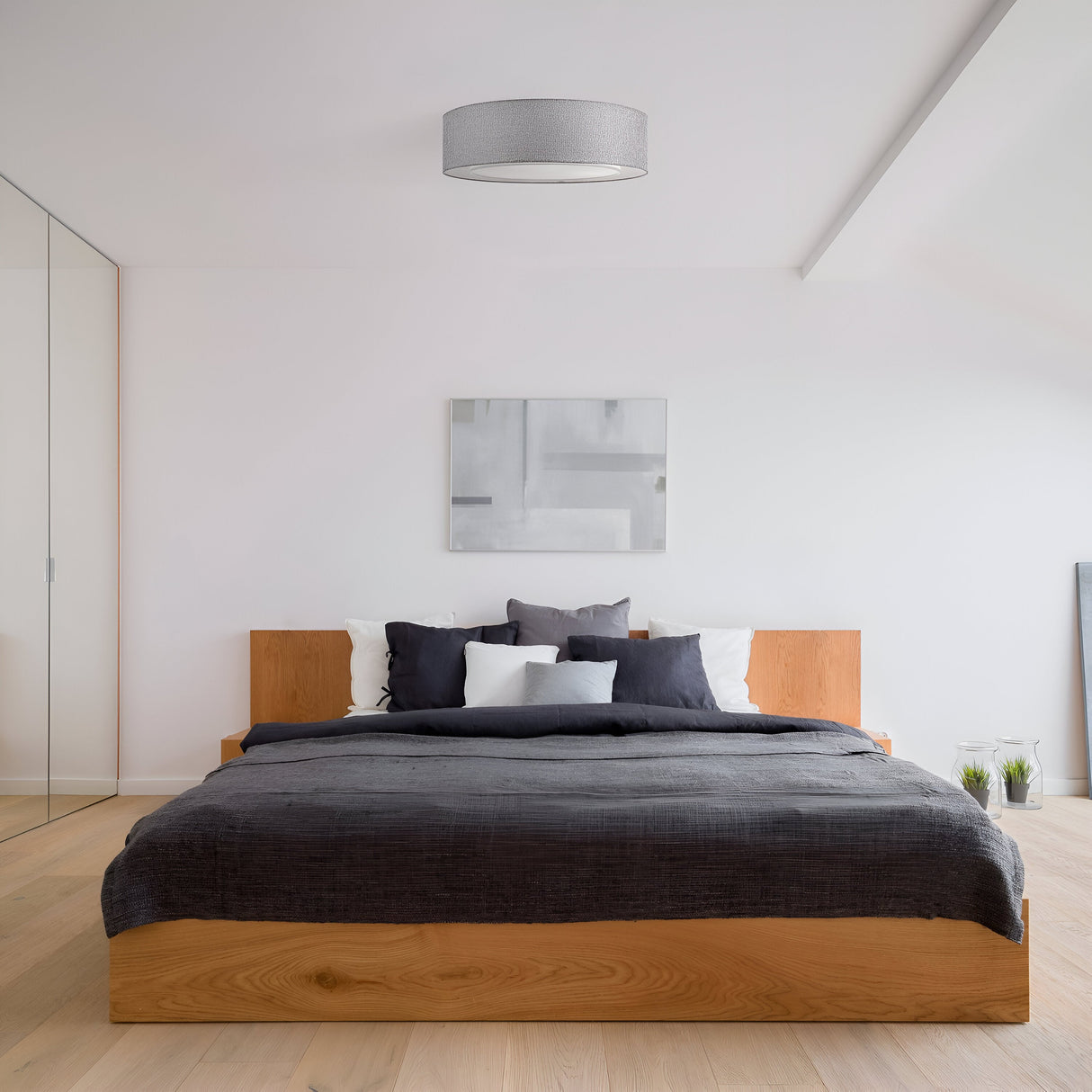 A minimalist bedroom showcases a large wooden bed with gray and white bedding, and a modern abstract painting on the wall above. The room features light wood flooring and is accented by the Bergamo 3 Light Flush in Chrome with Grey Shade, enhancing its contemporary design.
