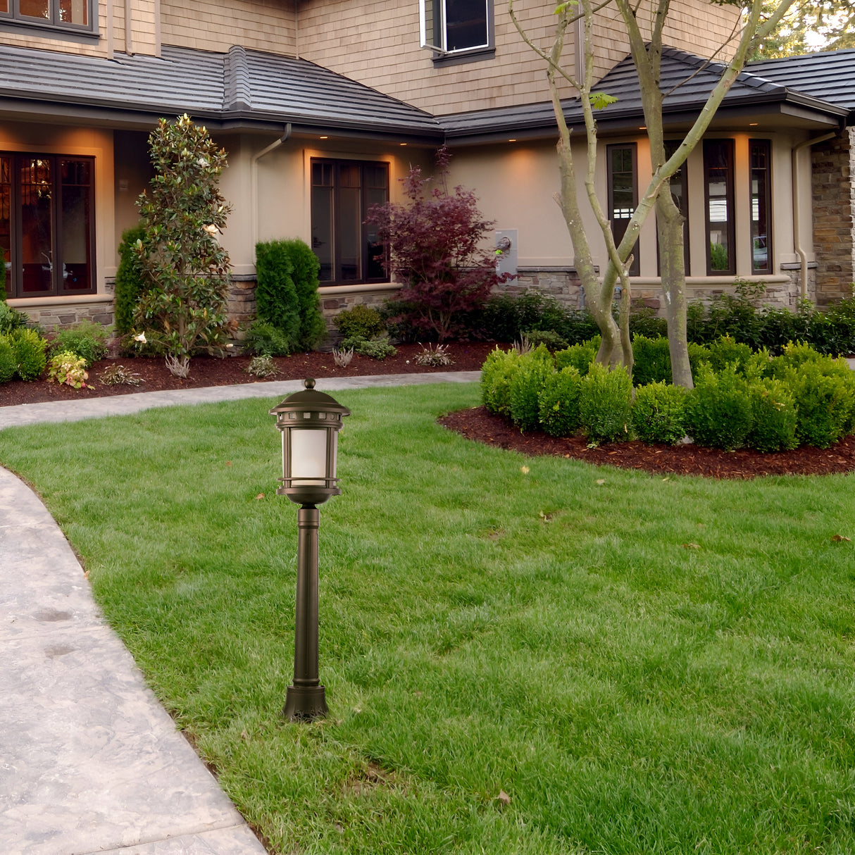 A suburban house with beige siding, dark roof, manicured lawn, and landscaped shrubs features a curved concrete path leading to the entrance. A vintage-look Salamanca 1 Light Pillar Lantern - Brown stands on the grass.